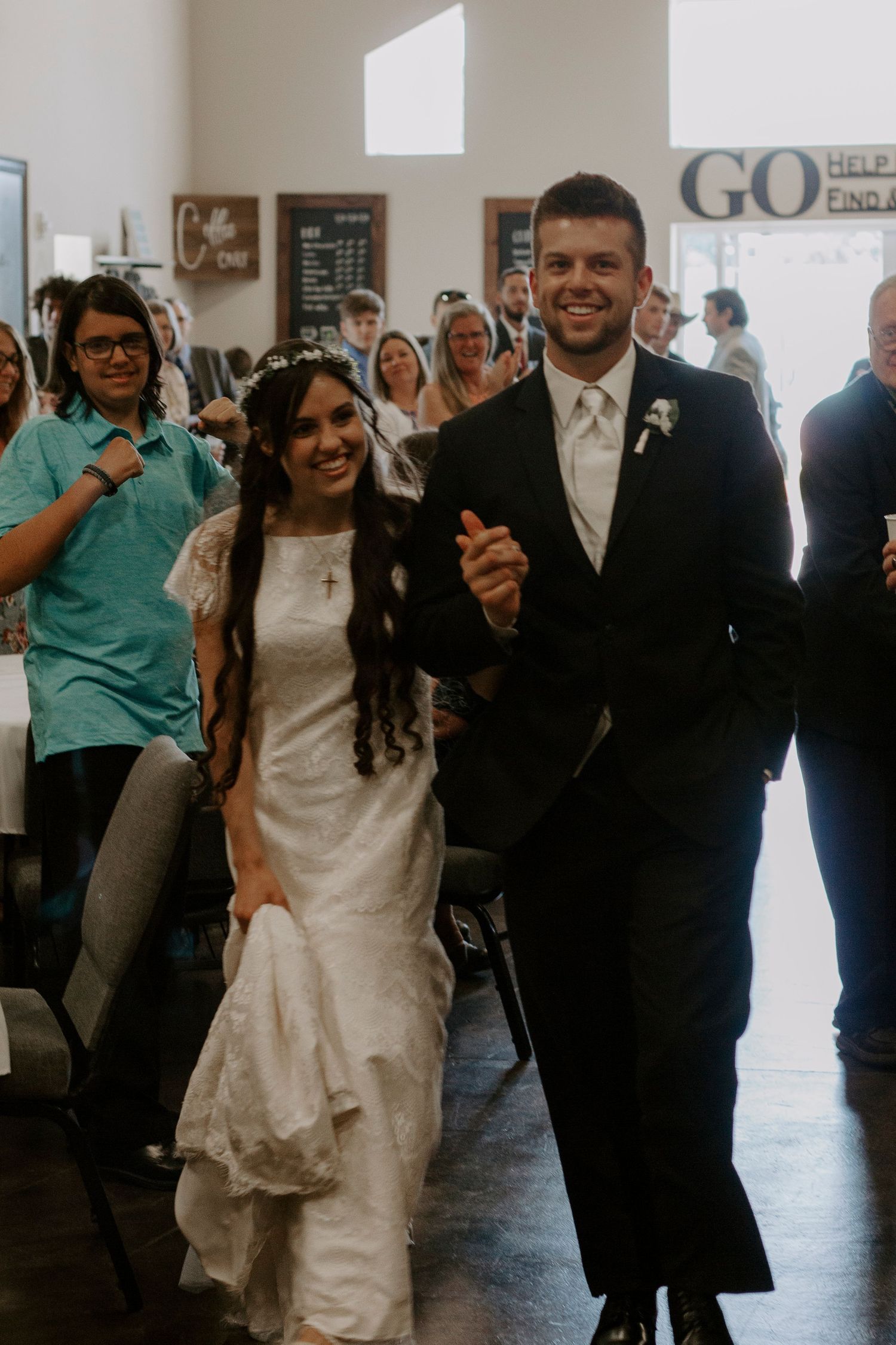 A smiling bride in a white dress and floral headband walks arm in arm with her groom through a crowd of wedding guests.