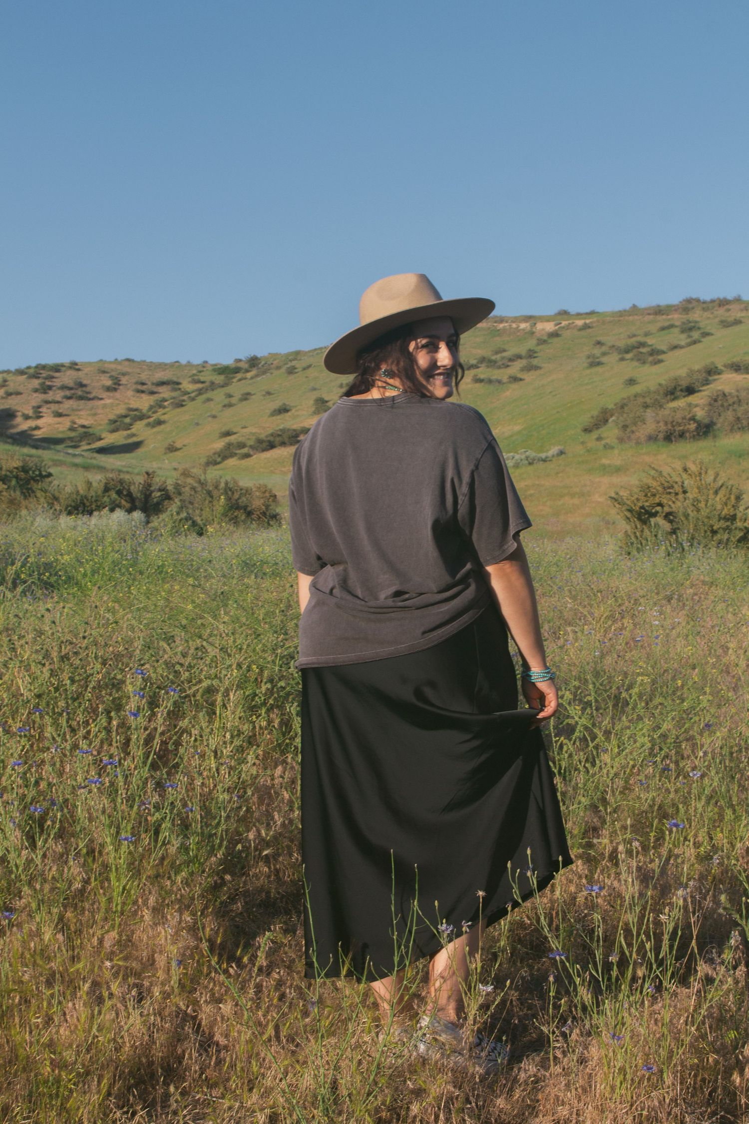 A woman in a tan hat, graphic tee and long black skirt looks back over her shoulder in a wildflower meadow.