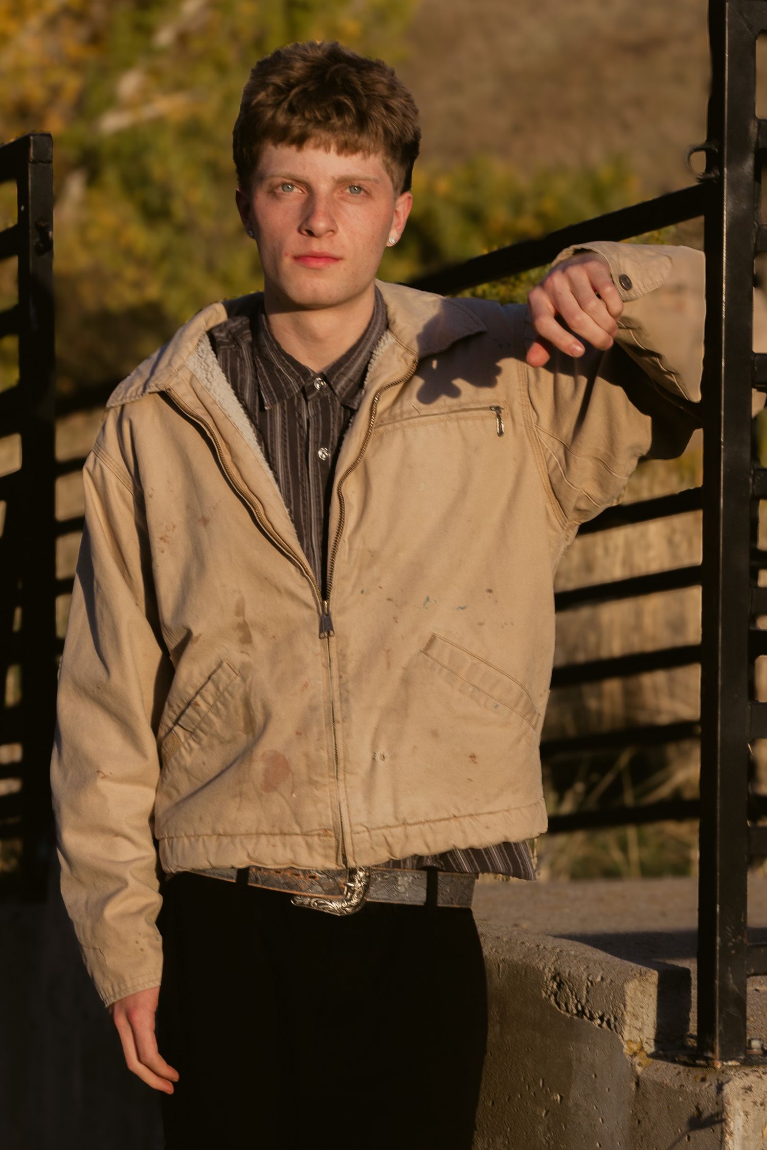 Young man in a dirty tan work jacket and dark pants leans an arm on a black metal railing in warm golden light.