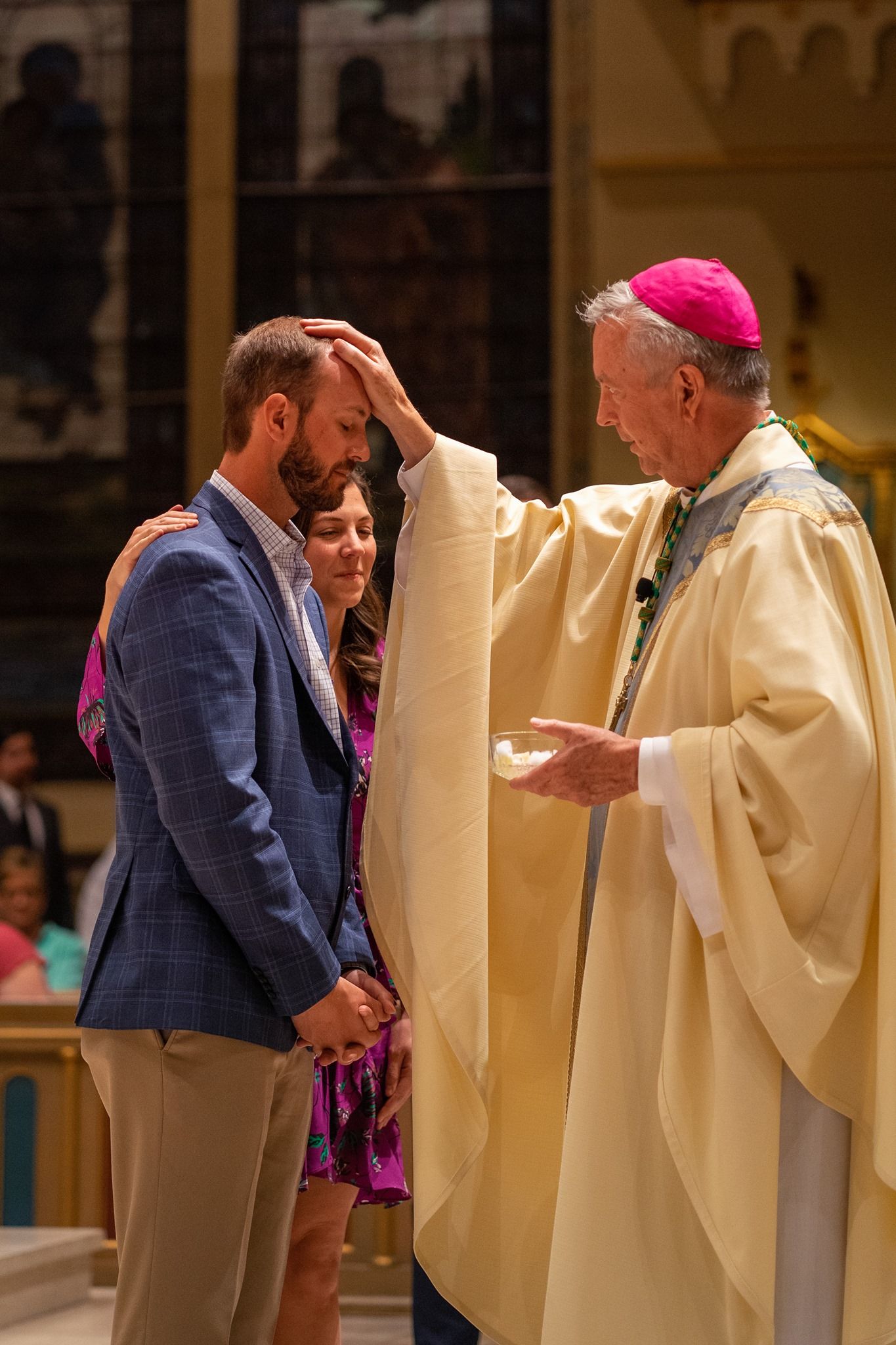A bishop in a pink zucchetto and cream vestments blesses a bearded man in a blue blazer as a woman stands beside him in a church.