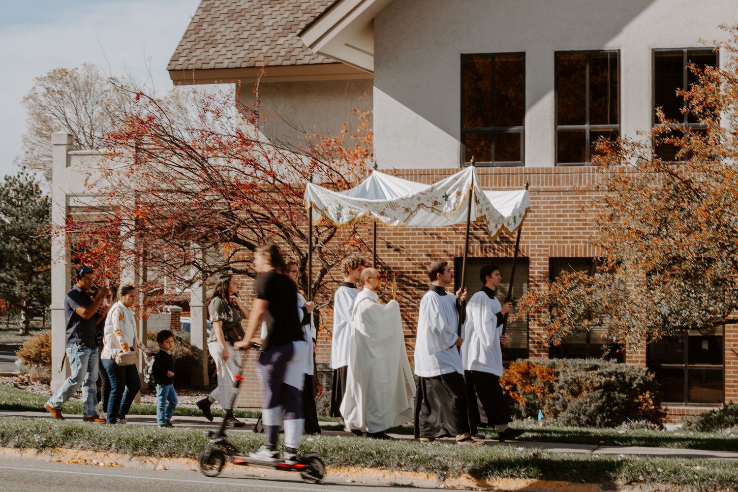 Clergy and parishioners walk in a Eucharistic procession under a white canopy past a brick church building on a sunny autumn day.