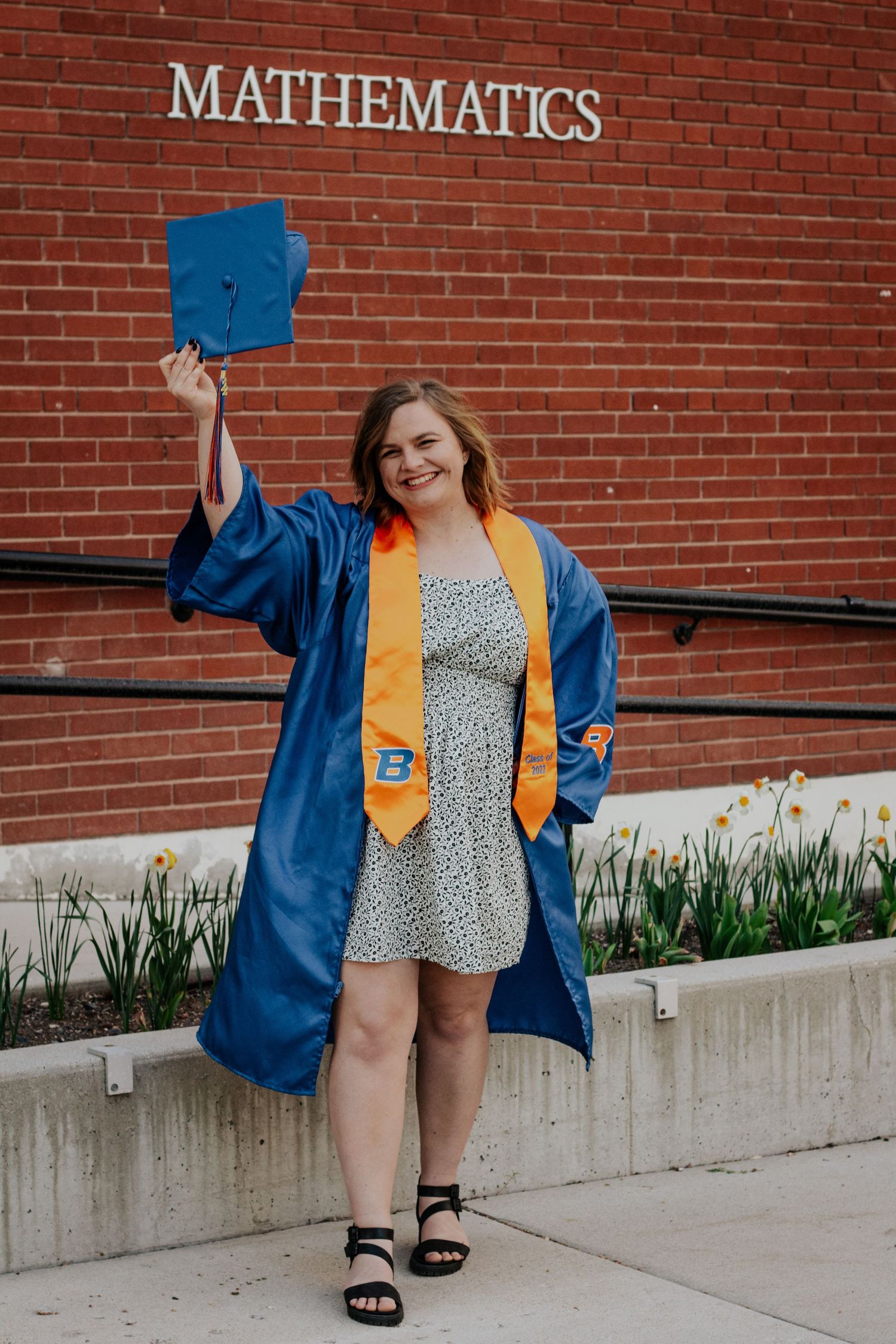 Graduate in a blue gown and orange stole raises her cap in celebration in front of a red brick Mathematics building at Boise State.