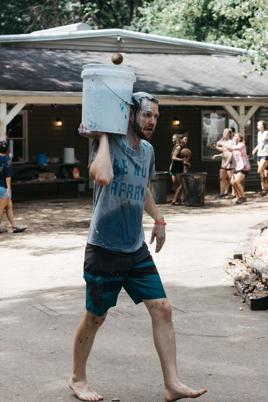 A barefoot man in a gray t-shirt and teal shorts carries a white bucket on his shoulder past a cabin at a summer camp.