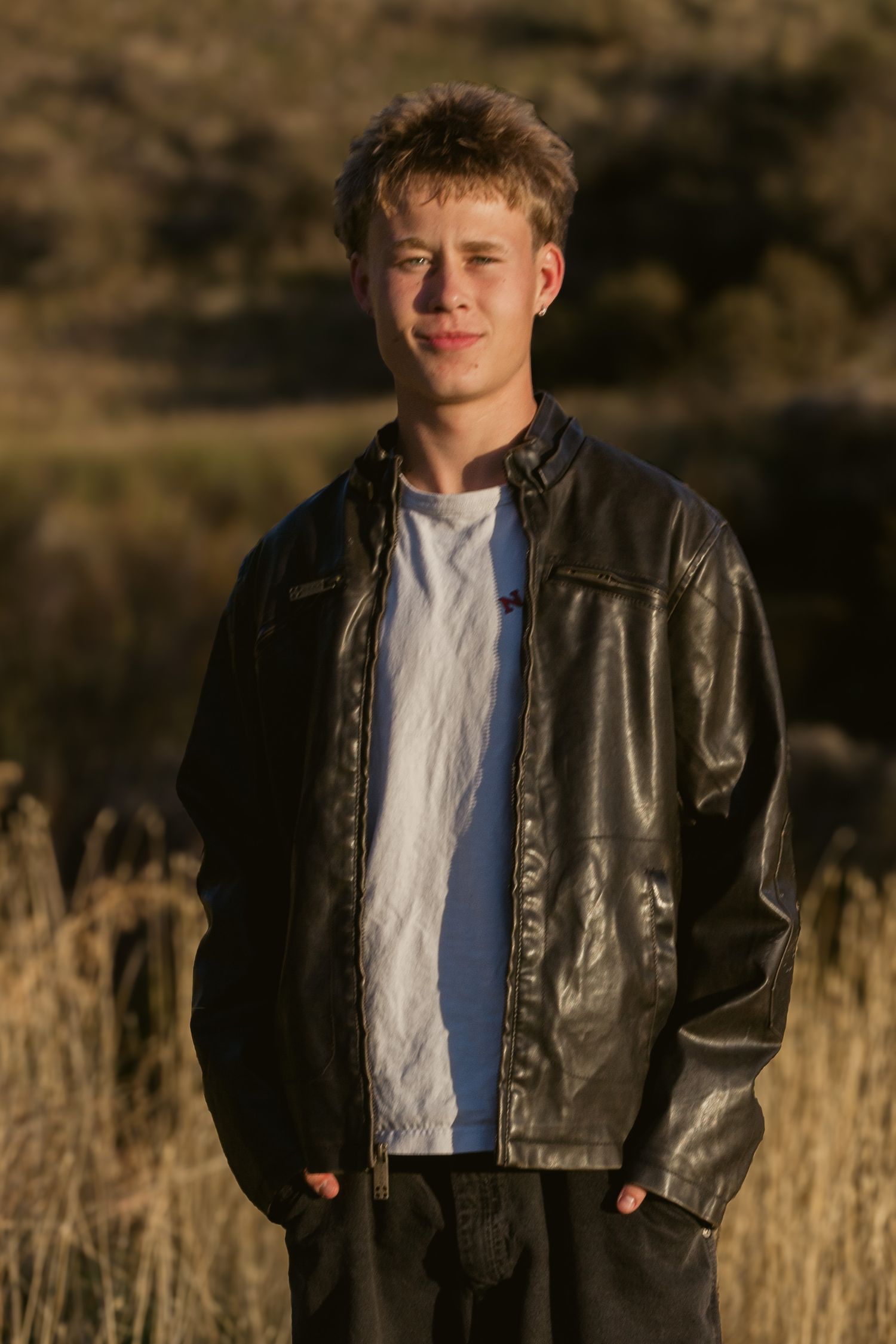 Young man in a black leather jacket and white t-shirt stands with hands in pockets in a golden grass field at sunset.