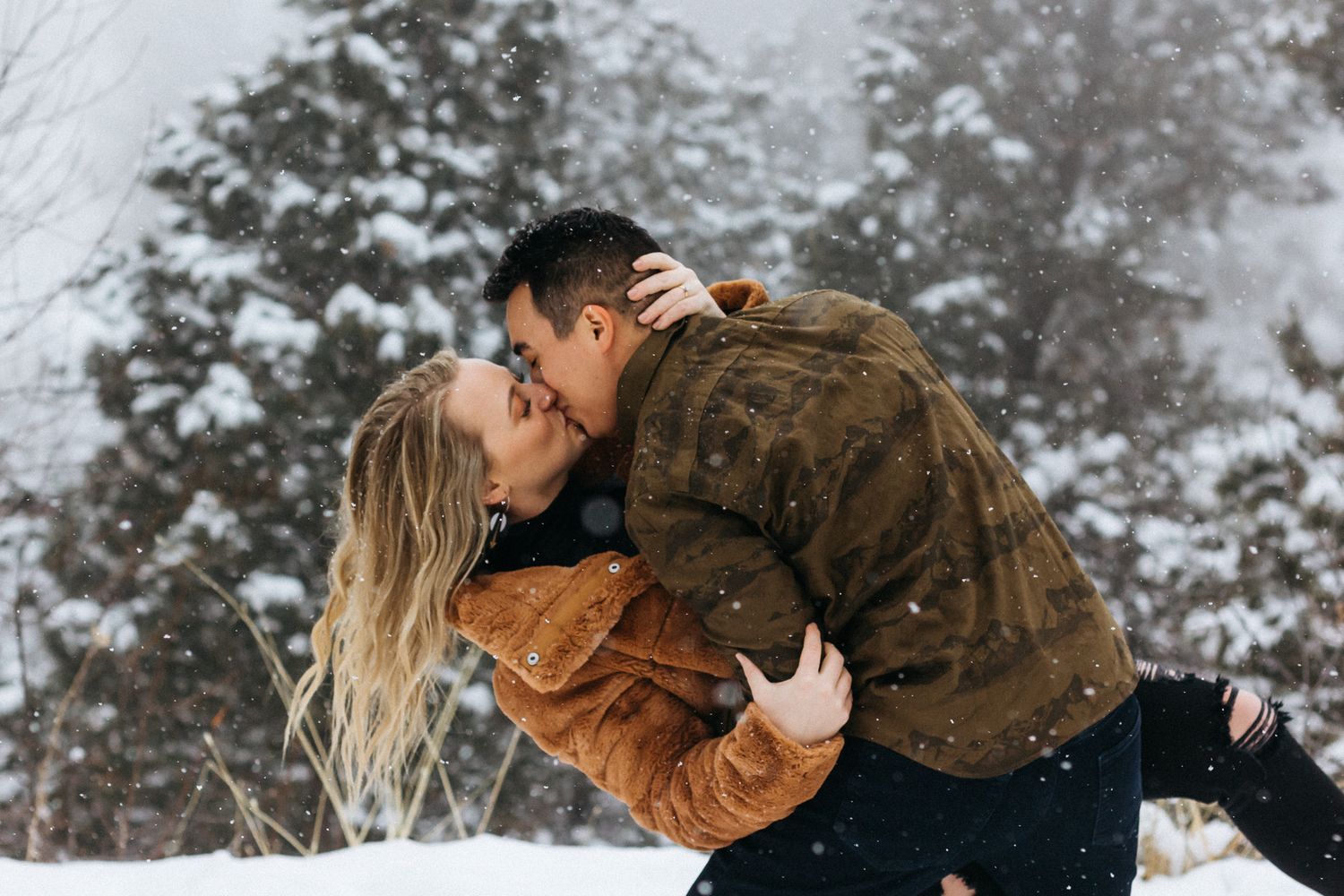 A couple shares a romantic dipped kiss in a snowy pine forest as the woman's long blonde hair falls back.