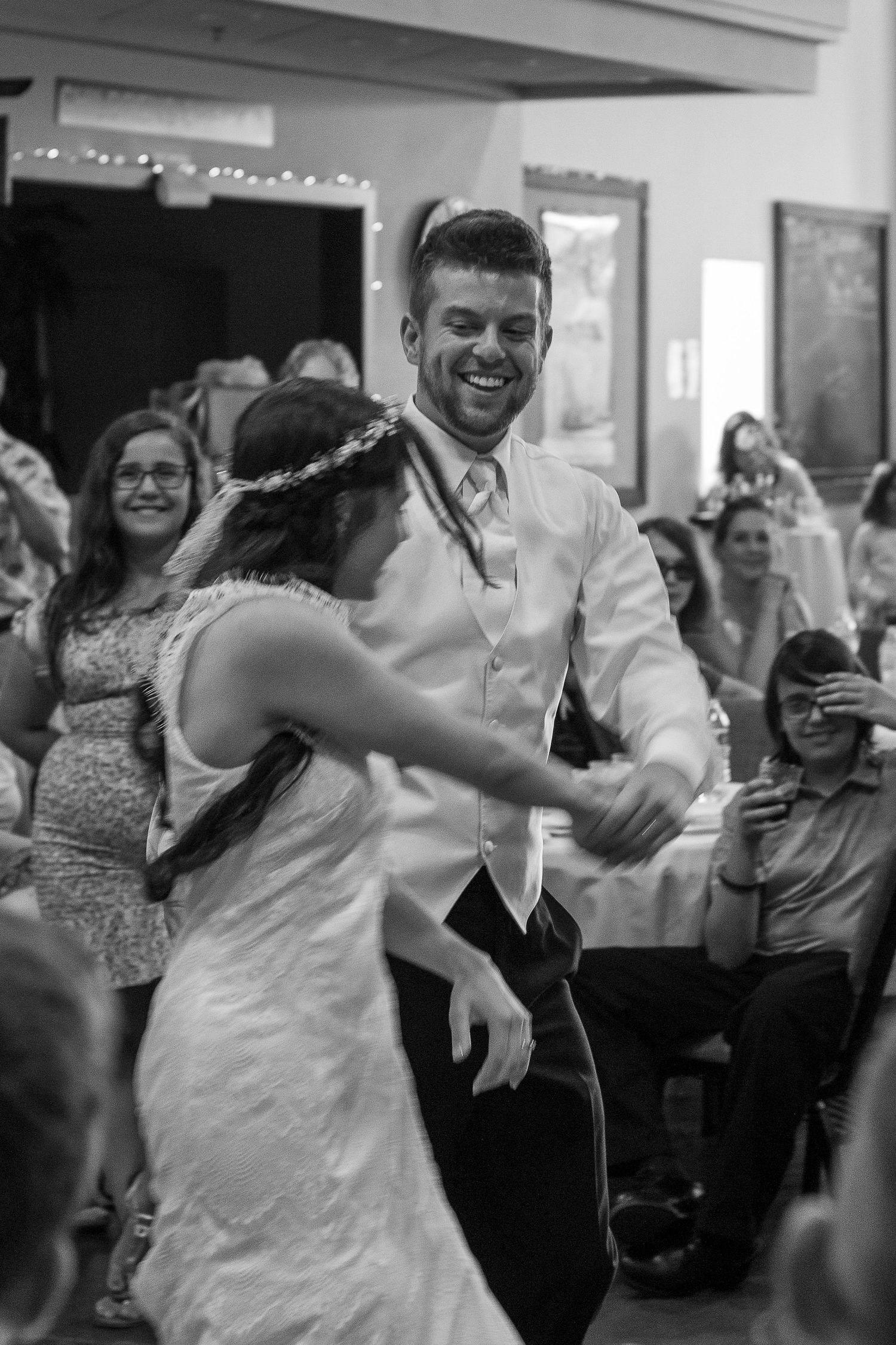 A laughing groom in a vest dances joyfully with his bride in a flower crown as guests watch in this black and white wedding reception photo.