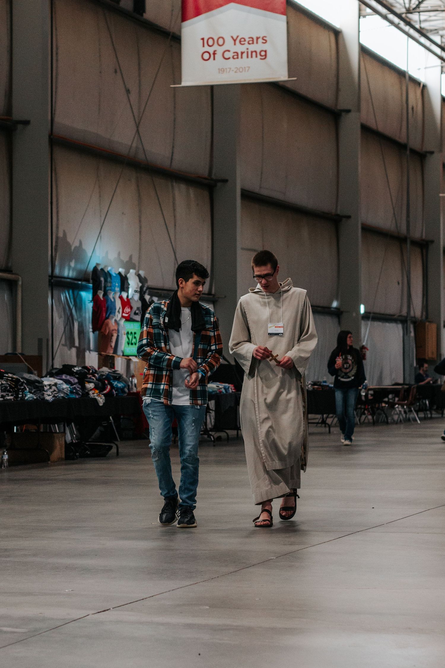 A teen in a plaid shirt walks and talks with a young man in a light religious habit under a 100 Years of Caring banner.