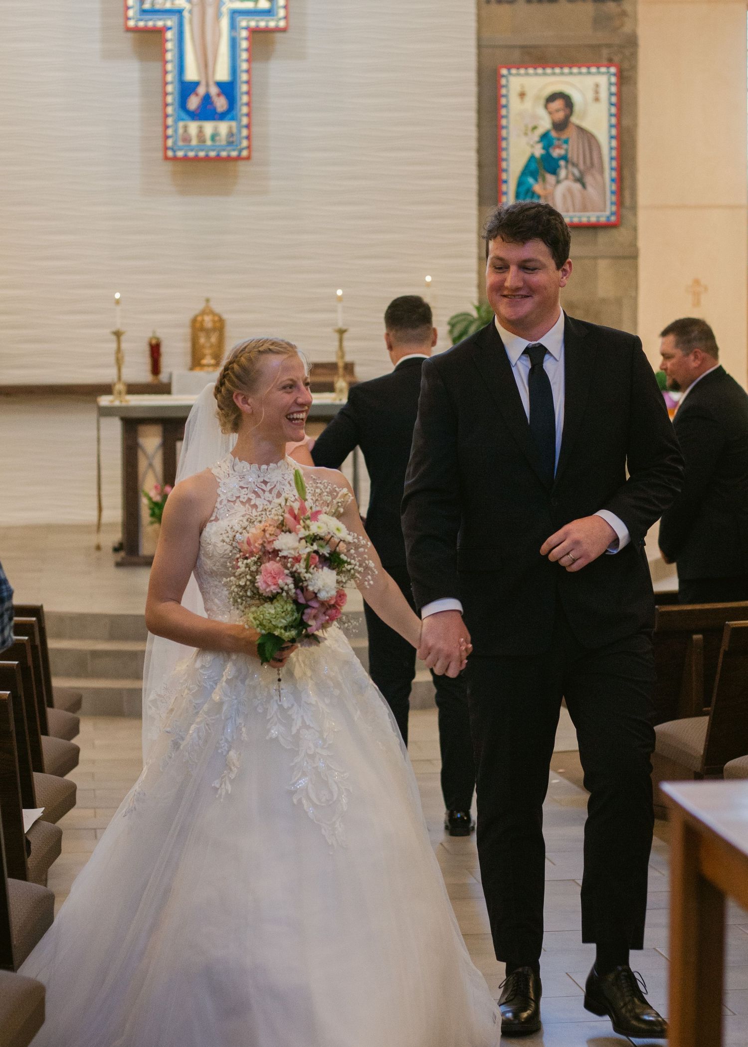 A smiling bride holding a pink floral bouquet walks hand in hand with her groom down the catholic church aisle after their wedding ceremony.