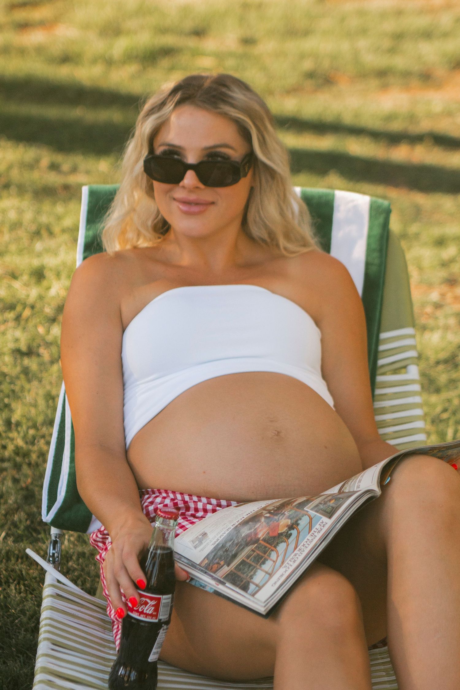 A smiling pregnant woman in black sunglasses and a white bandeau top lounges on a striped lawn chair holding a magazine and a cola bottle.