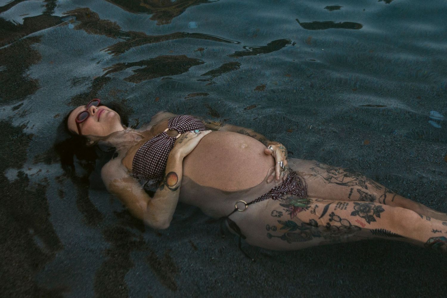 Pregnant woman in a brown bikini floats peacefully on her back in dark pool water, hands resting on her baby bump.