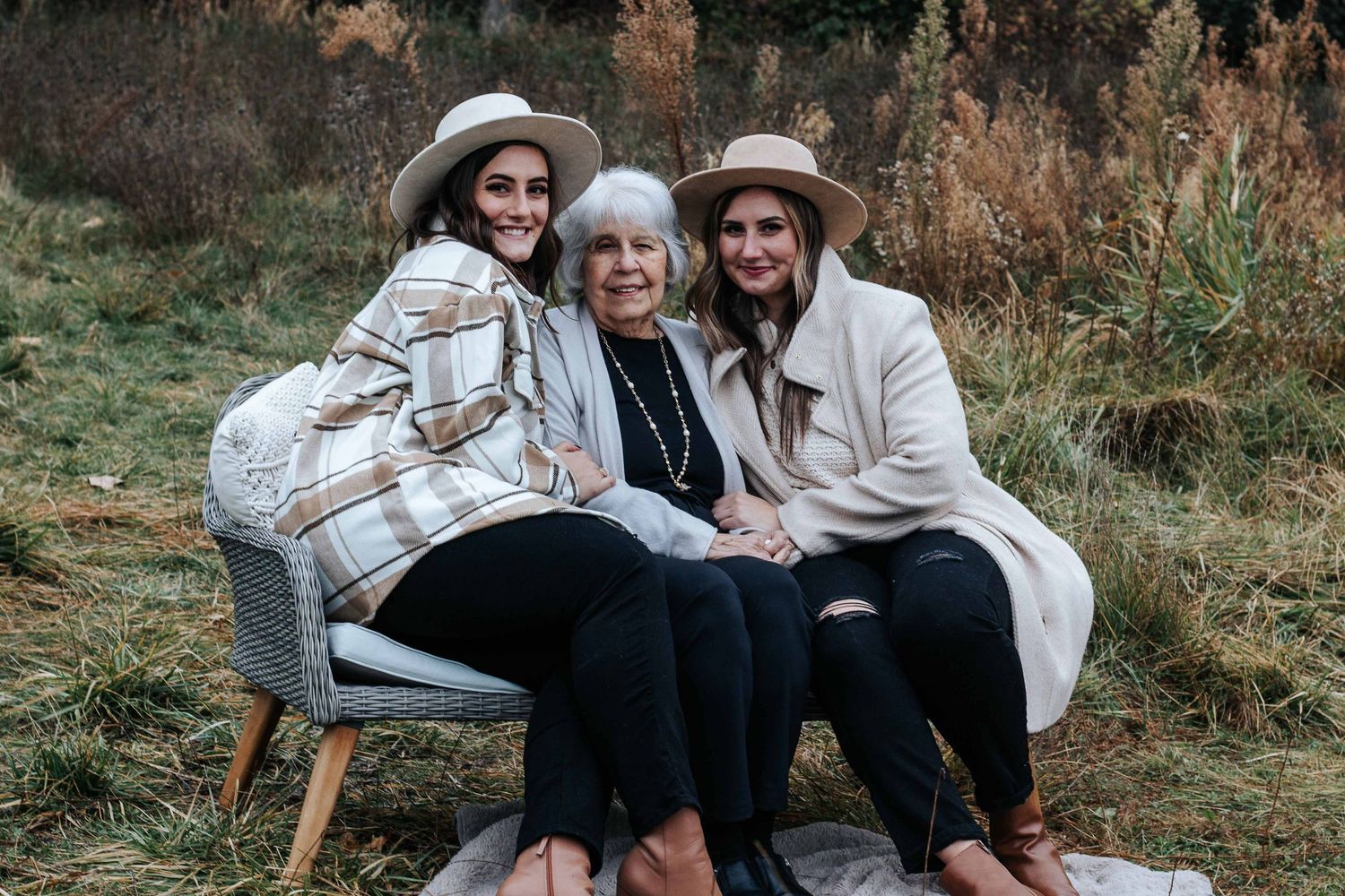 Two young women in wide-brim hats lean in close to a smiling older woman seated on a bench in an autumn field.