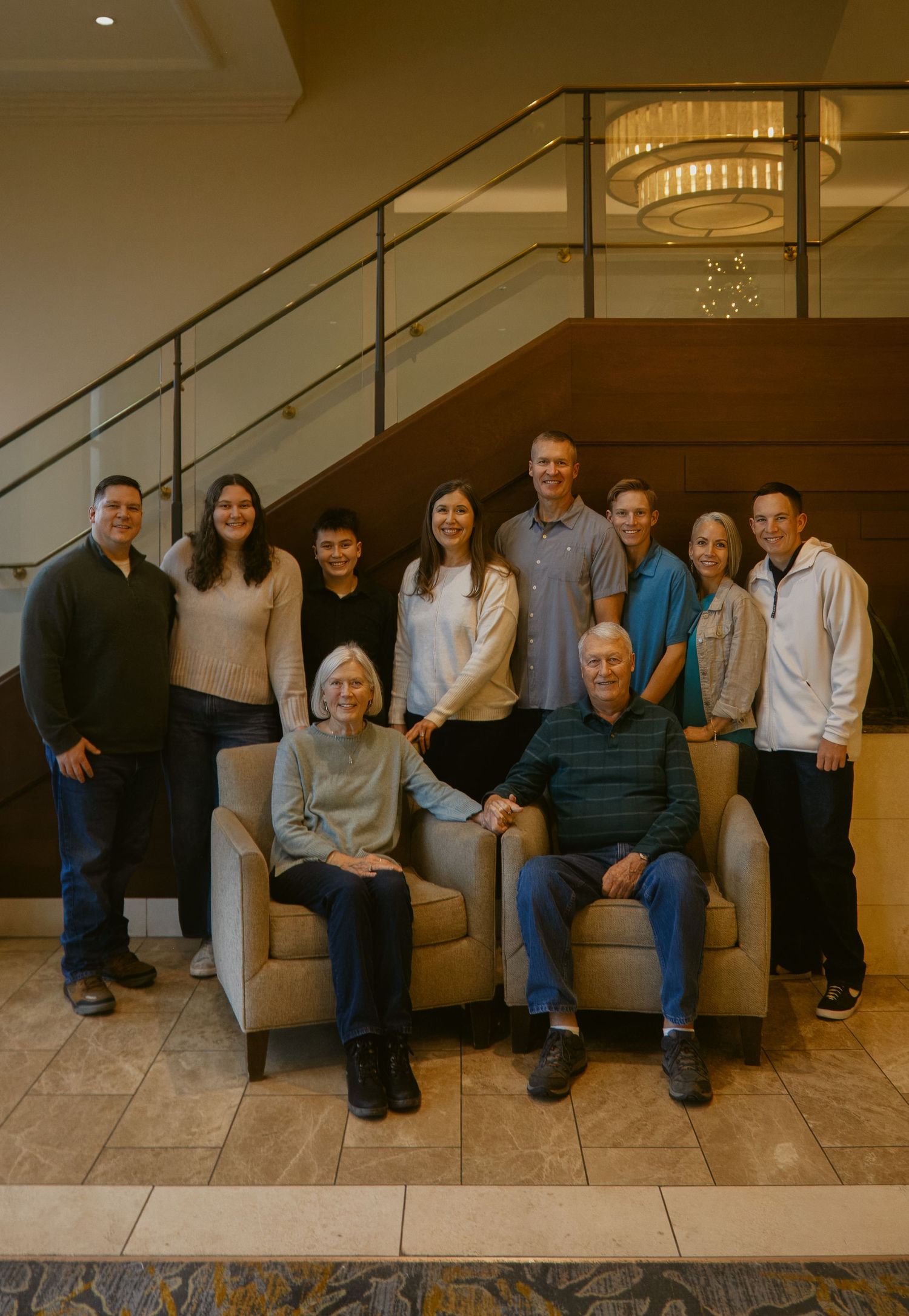 Multigenerational family of ten posing together by a staircase in a lobby, with two seniors seated in front.