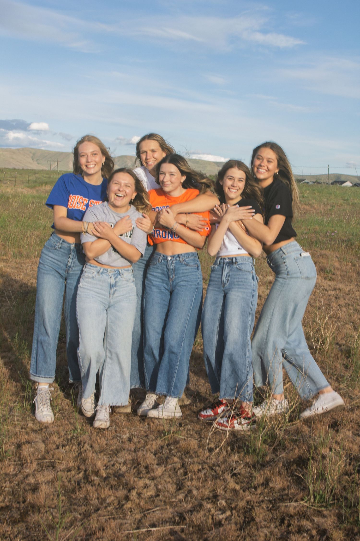 Six smiling friends in jeans and school-spirit tops hug close together in a grassy field under a blue sky with scattered clouds.