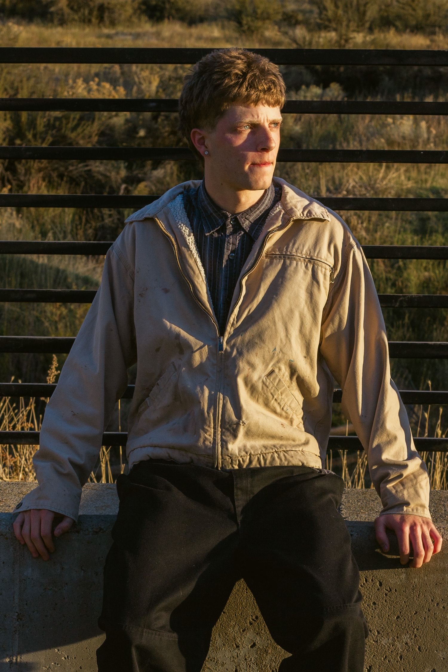 Young man in a tan work jacket sits on a concrete ledge in golden sunlight, looking off to the side beside a black railing.