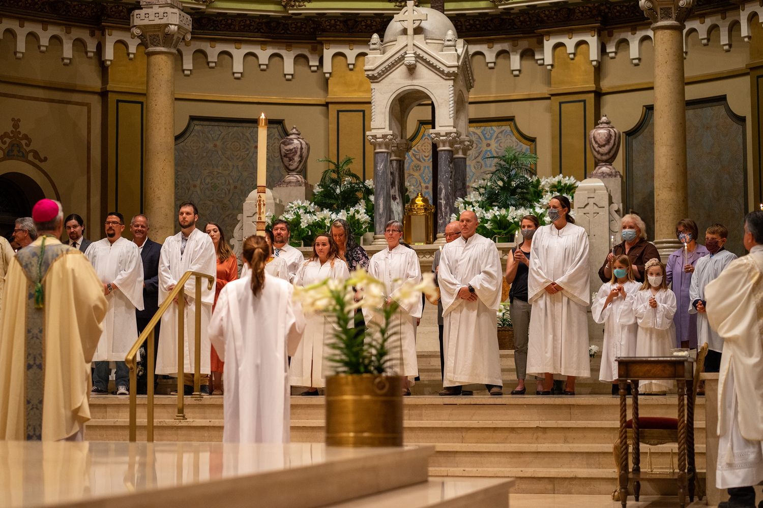 Clergy, altar servers, and worshippers in white robes gather near the altar of an ornate church with white flowers and candles.
