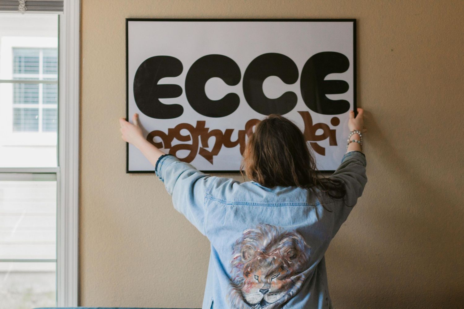 Woman in a denim jacket hangs a large ECCE sign with decorative lettering on a beige wall beside a window.