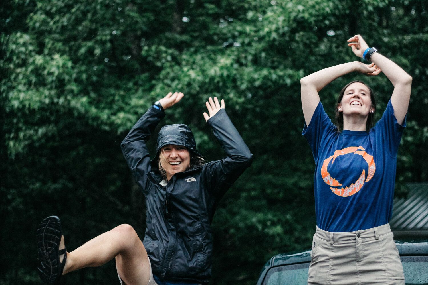 Two people in rain gear and a blue t-shirt raise their arms and laugh playfully outdoors in front of lush green trees.