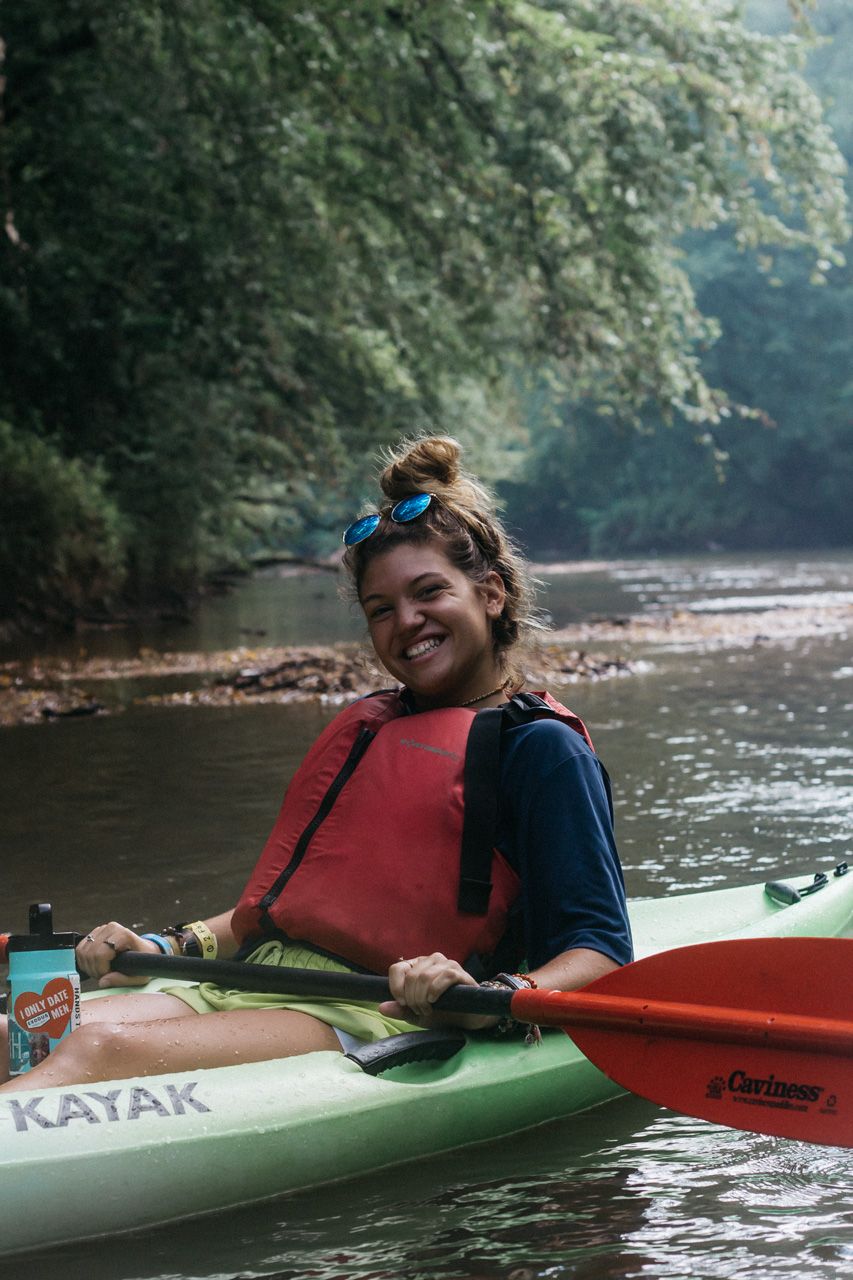 A smiling woman in a red life vest and sunglasses on her head holds a paddle while sitting in a green kayak on a calm river.