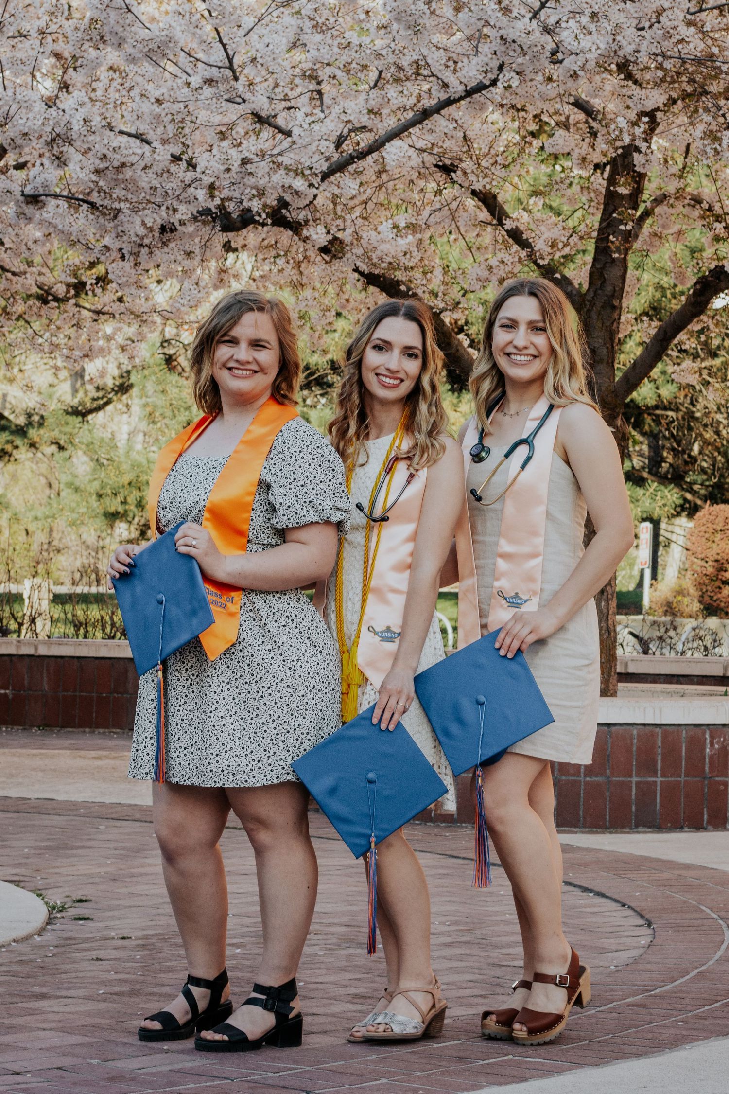 Three smiling female graduates hold blue caps and wear stoles while posing under a blooming cherry tree on a brick campus walkway.