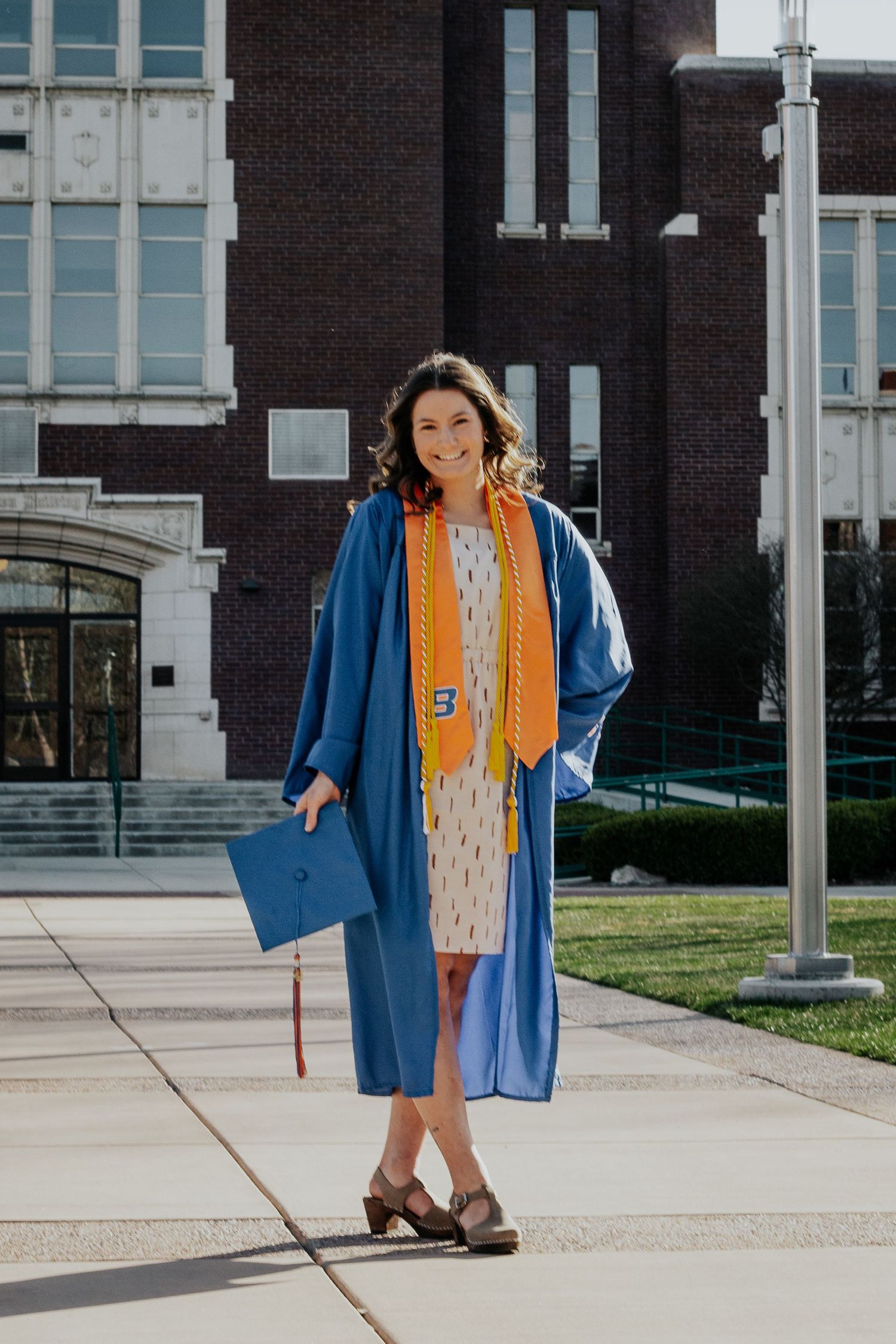 A smiling graduate in a blue gown and orange stole holds her cap while standing on a campus walkway by a brick building.