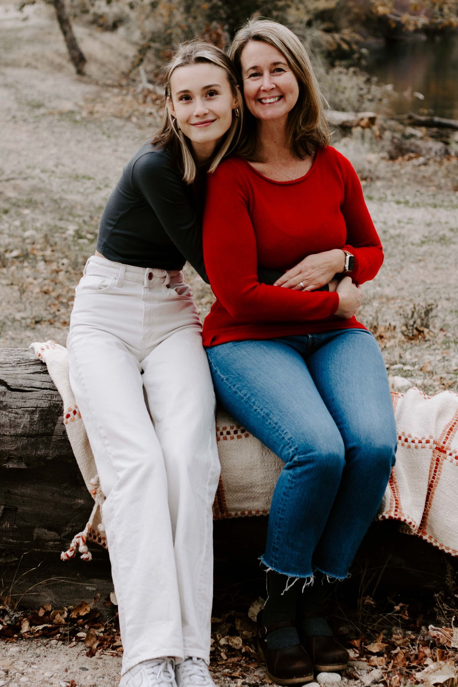 Smiling mother and teenage daughter sit close together on a log draped with a blanket in an autumn setting.