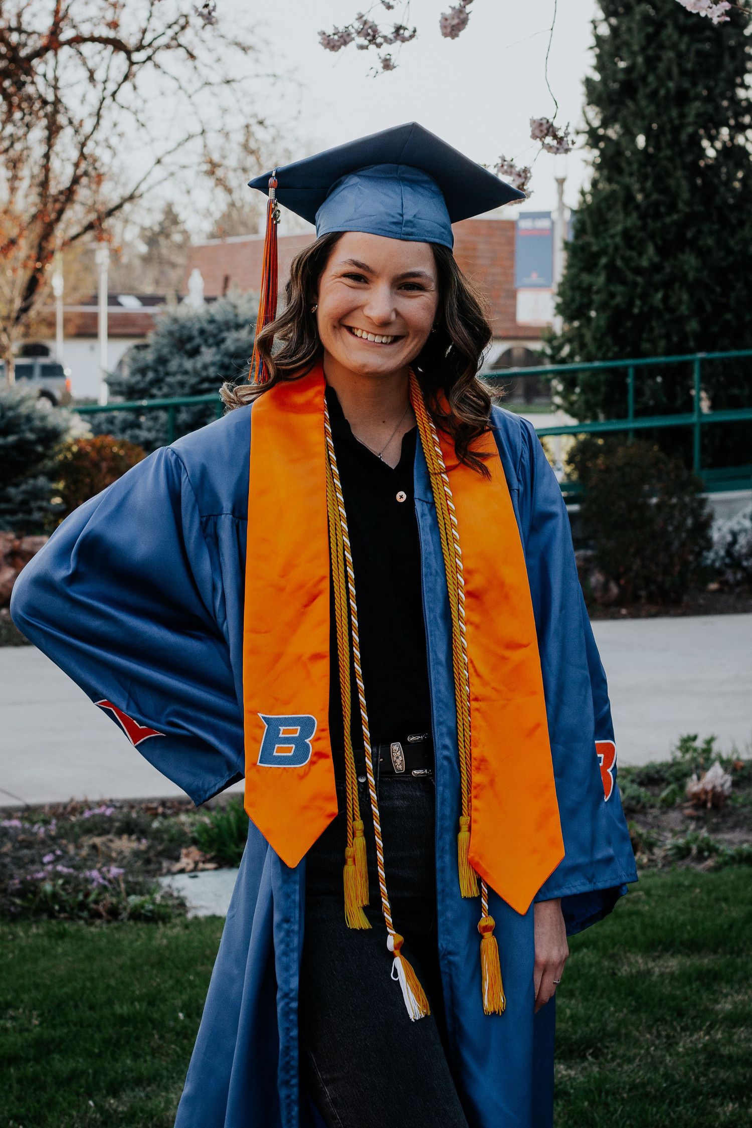 A smiling graduate in a blue cap and gown with an orange stole and gold cords poses outdoors on a spring day.
