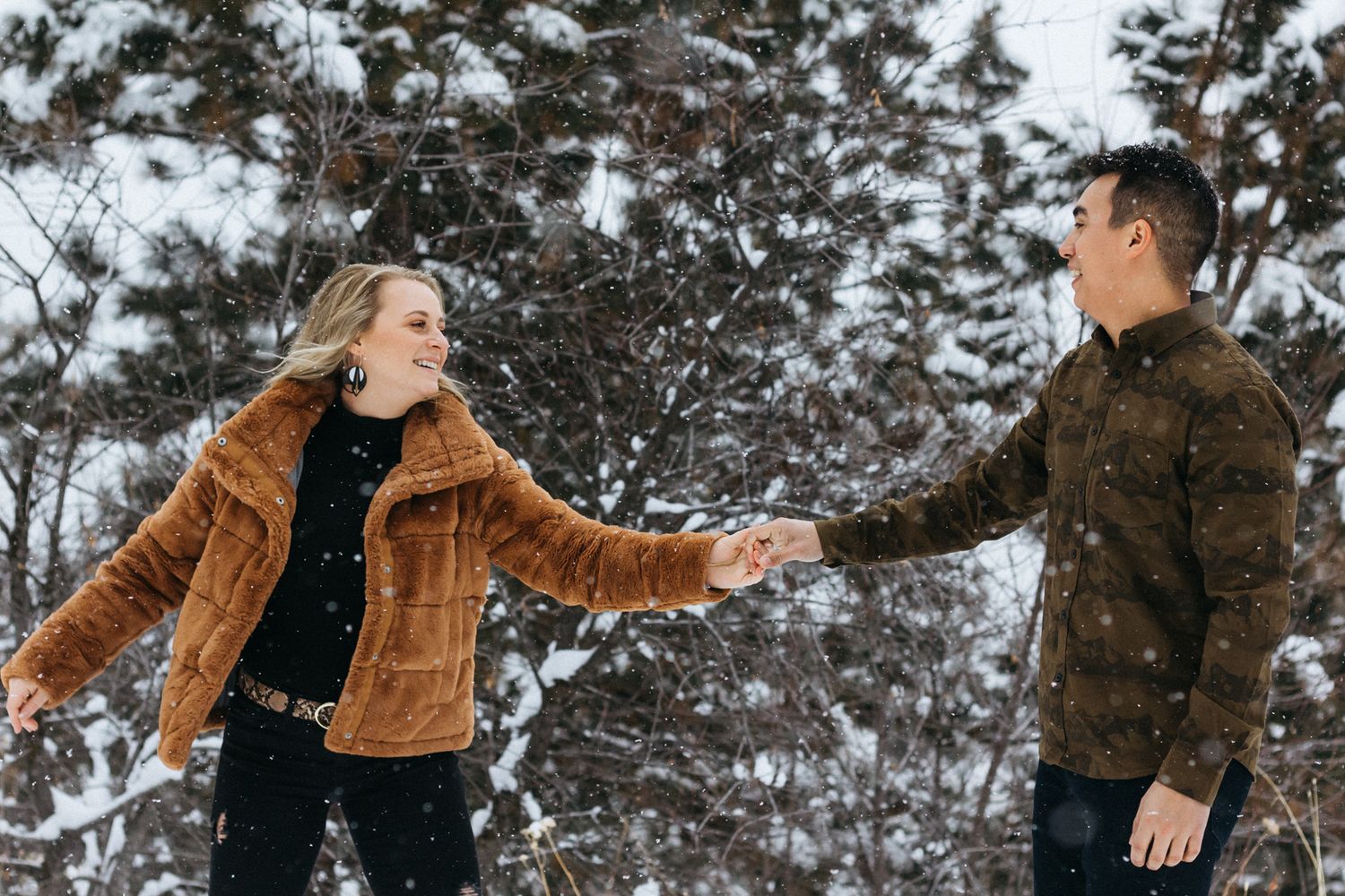 A woman in a fur-trimmed coat laughs and holds hands with a man in a camo jacket among snow-covered evergreen trees.