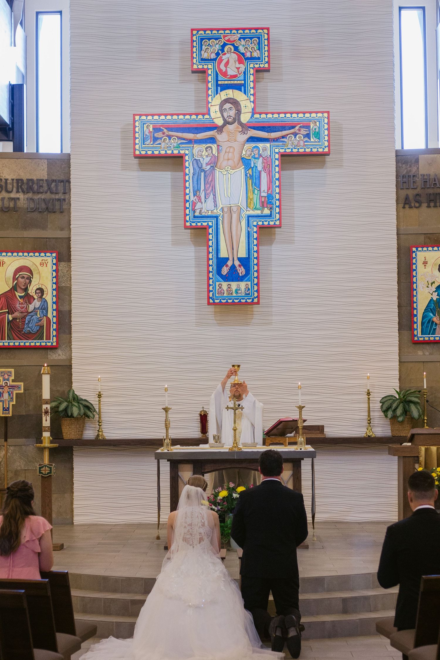 A bride and groom kneel before a priest at the altar beneath a colorful San Damiano crucifix during a Catholic wedding ceremony.
