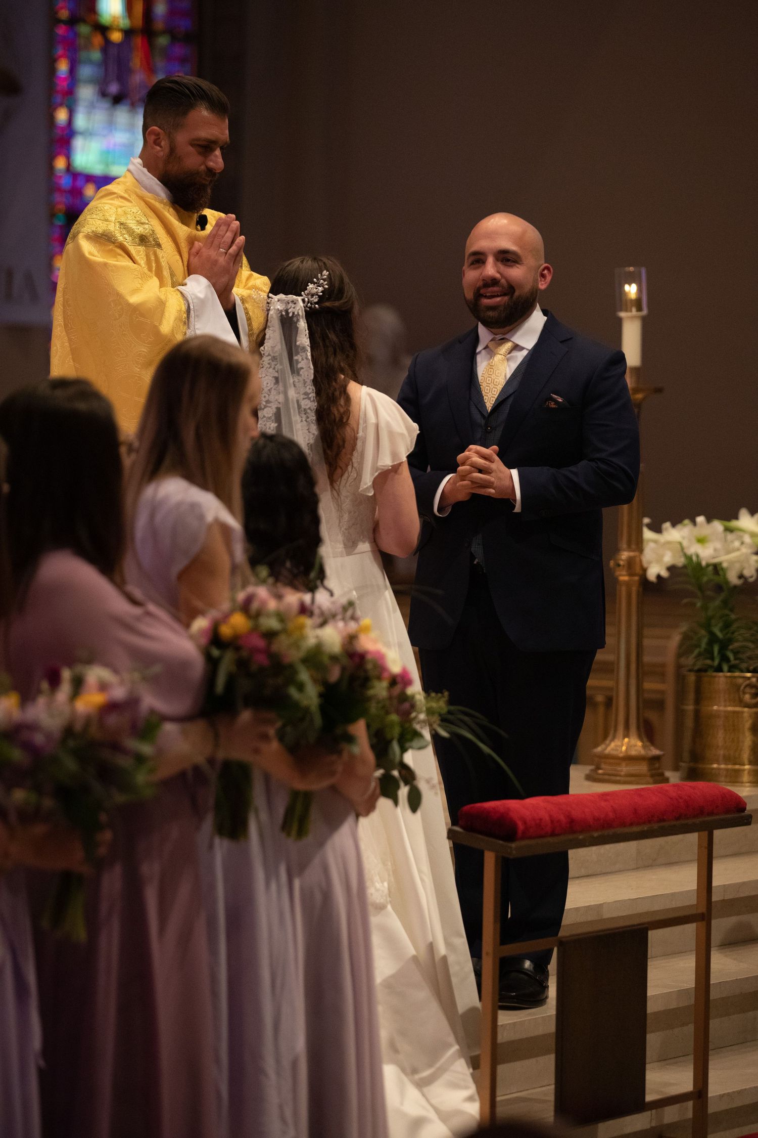 A groom smiles and folds his hands as a priest in yellow vestments officiates while bridesmaids in blush dresses hold bouquets.