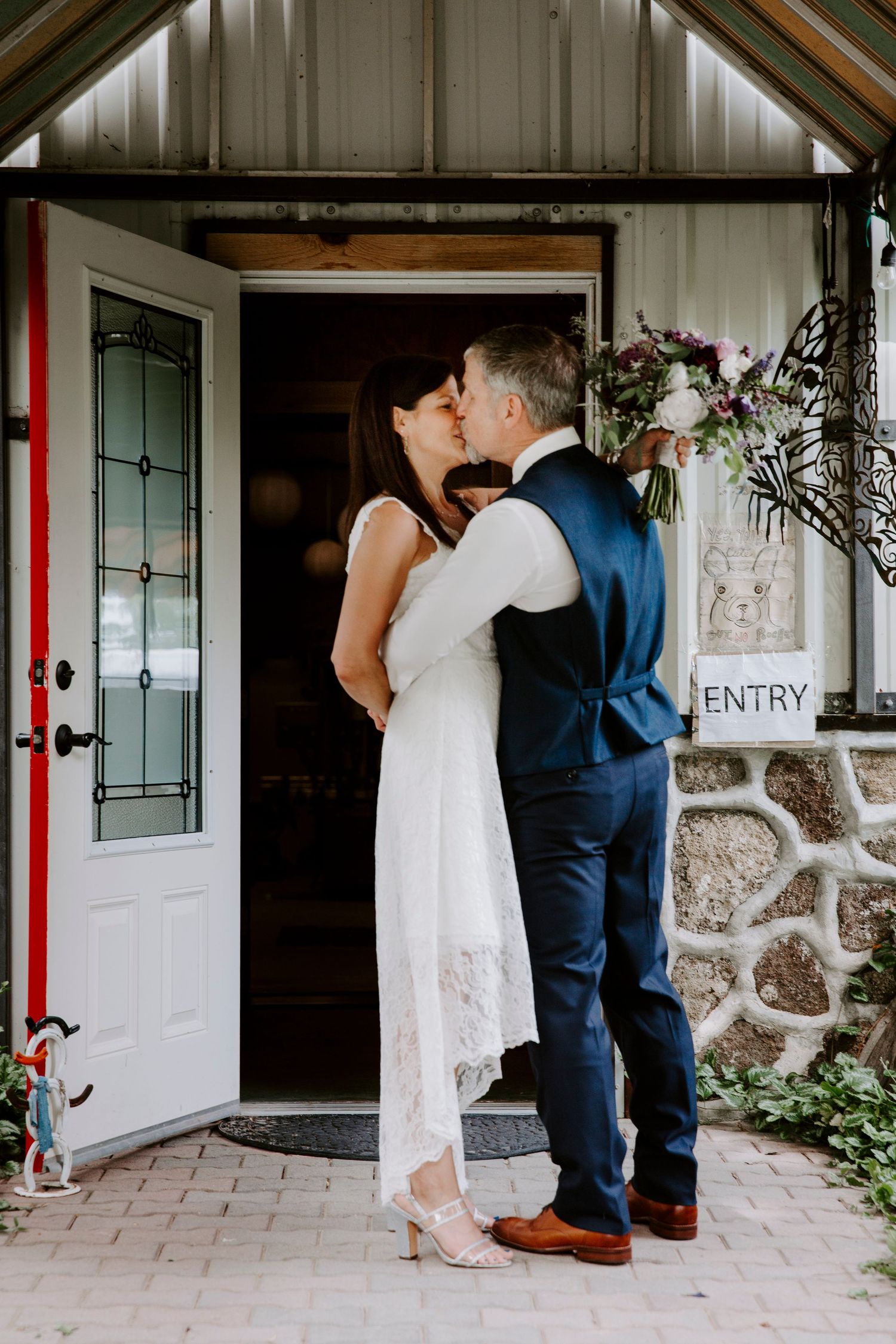 A bride in a white lace dress and a groom in a navy vest share a kiss in front of a white door beside an entry sign.