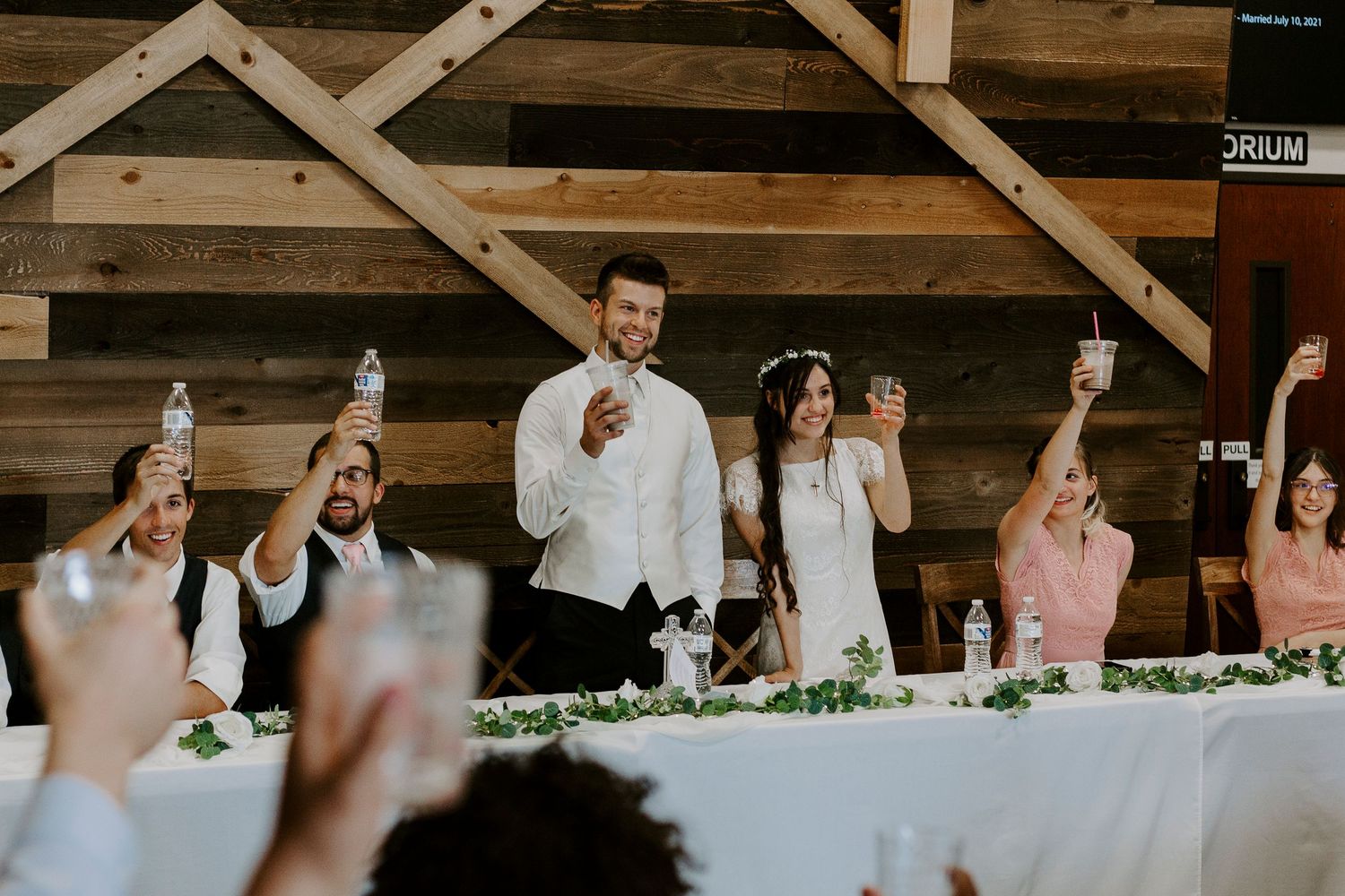 Wedding guests raise glasses in a toast at a long reception table decorated with greenery inside a rustic wooden barn venue.