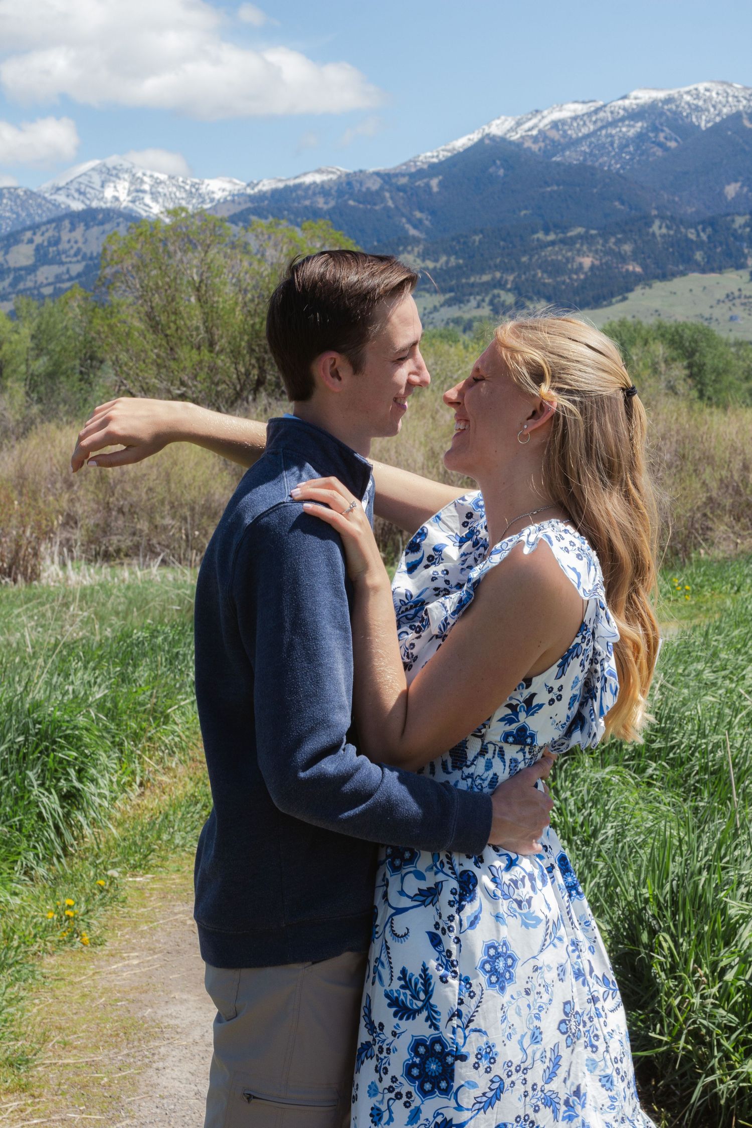 Couple embraces and smiles in a grassy field with snow-capped mountains and blue sky in the background.