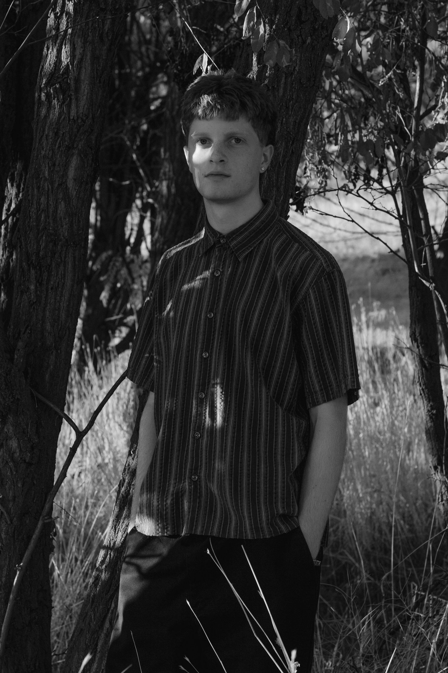 Black and white portrait of a young man in a striped short-sleeve shirt standing among trees and tall grass.