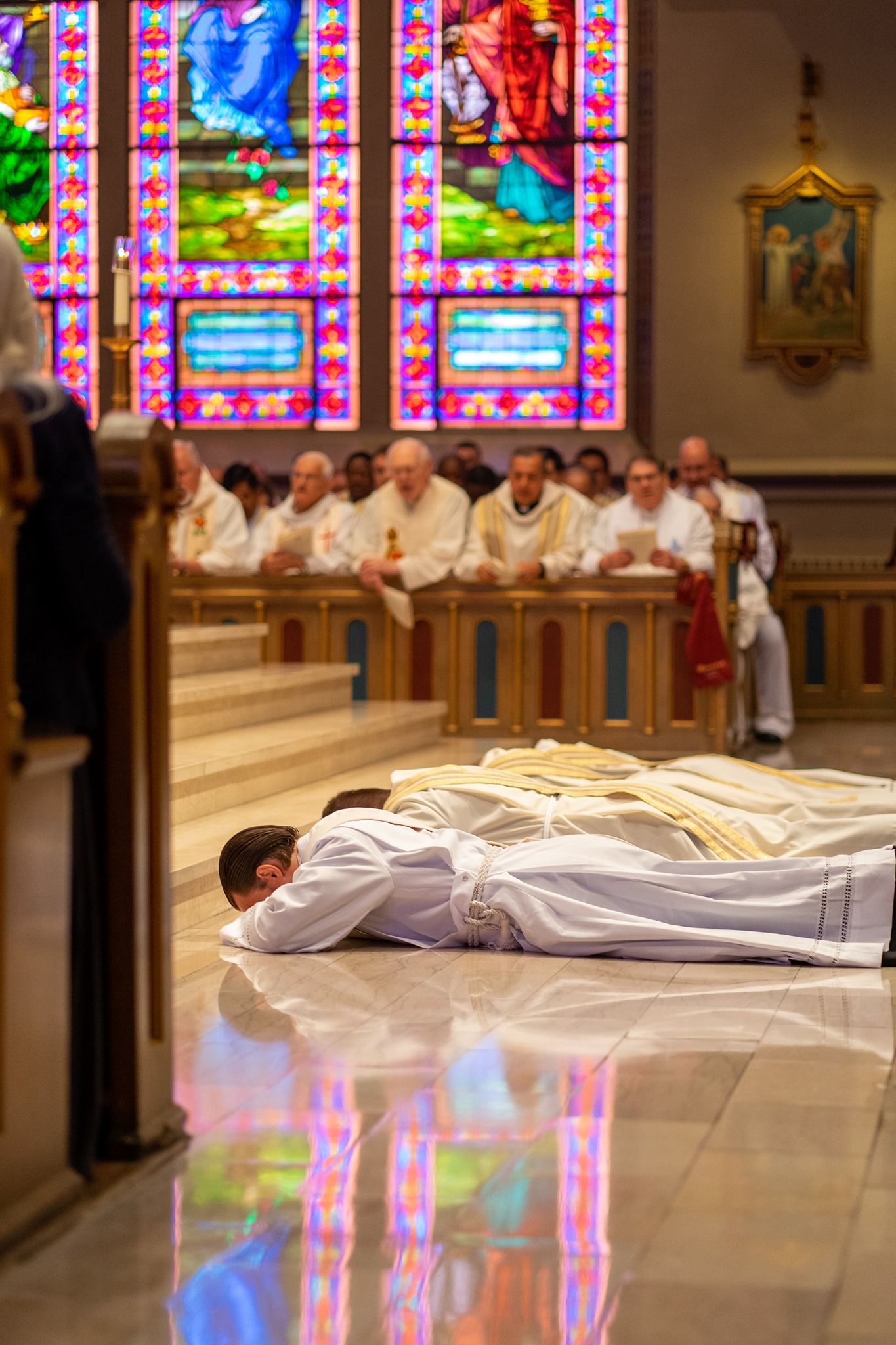 An ordination candidate in white lies prostrate on a shiny church floor as clergy sit behind a wooden rail beneath stained glass windows.