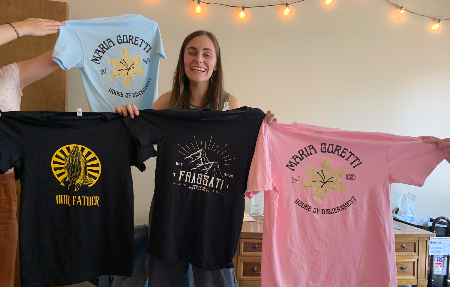 Smiling young woman holds up four colorful t-shirts, including a pink Maria Goretti shirt and a black Frassati shirt, in a cozy room.
