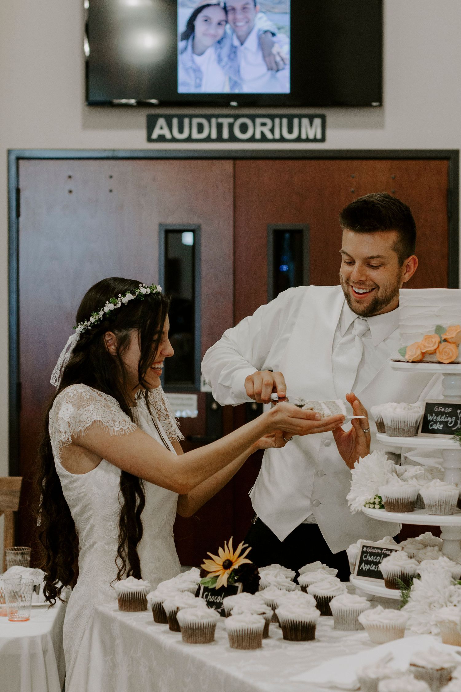 A laughing bride and groom cut into a table of cupcakes together at their wedding reception in front of an auditorium door.