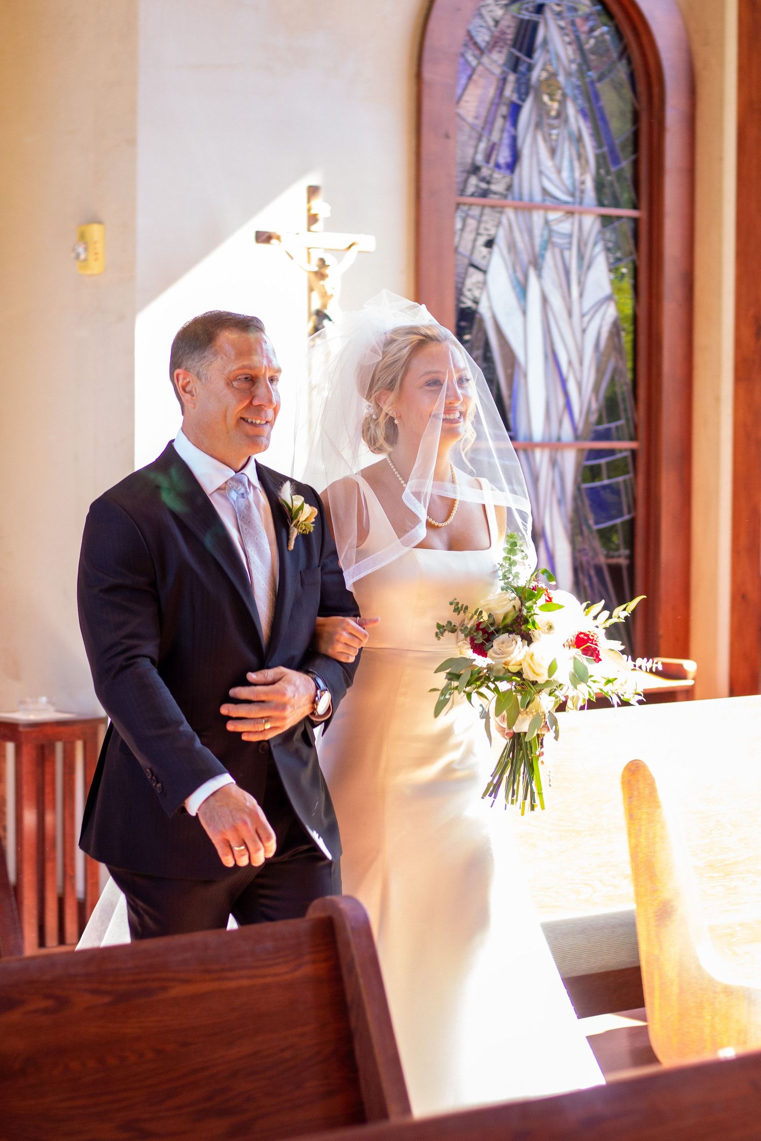 A smiling bride in a veil and white gown holds a bouquet while walking arm in arm with a man in a suit down a catholic church aisle.