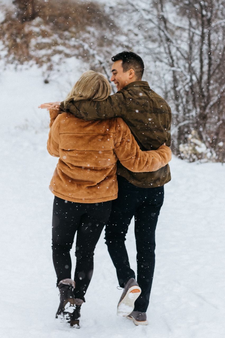 A couple in cozy winter jackets walk arm in arm and laugh along a snowy forest trail while snowflakes fall.