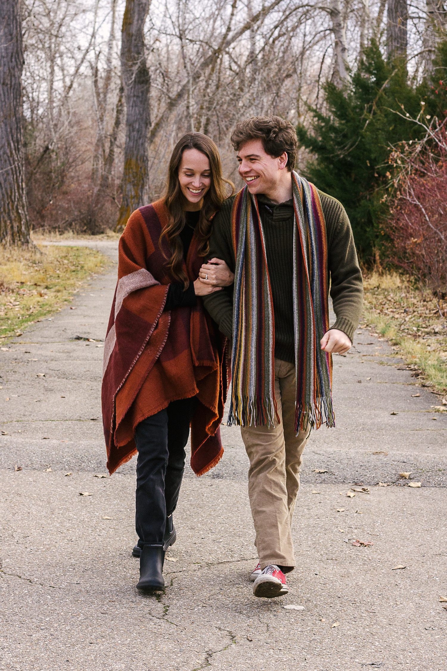 Happy couple walks arm in arm along a paved path through autumn woods, wearing scarves and a rust-colored wrap.