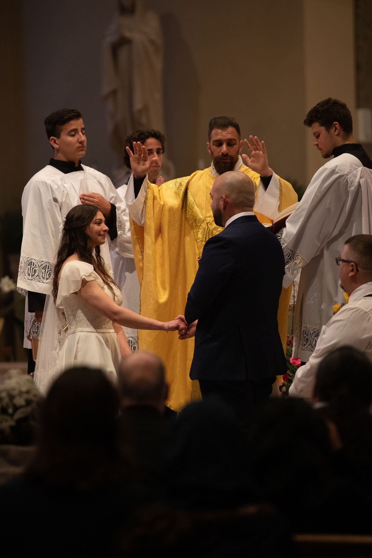 A priest in golden vestments blesses a bride and groom holding hands as altar servers look on during a church wedding.
