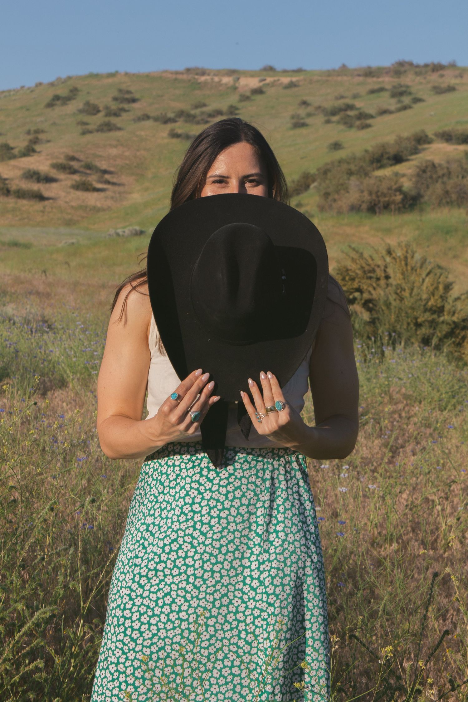 Woman in a green floral skirt holds a black cowboy hat over her face, peeking over the brim in a field of wild grasses.