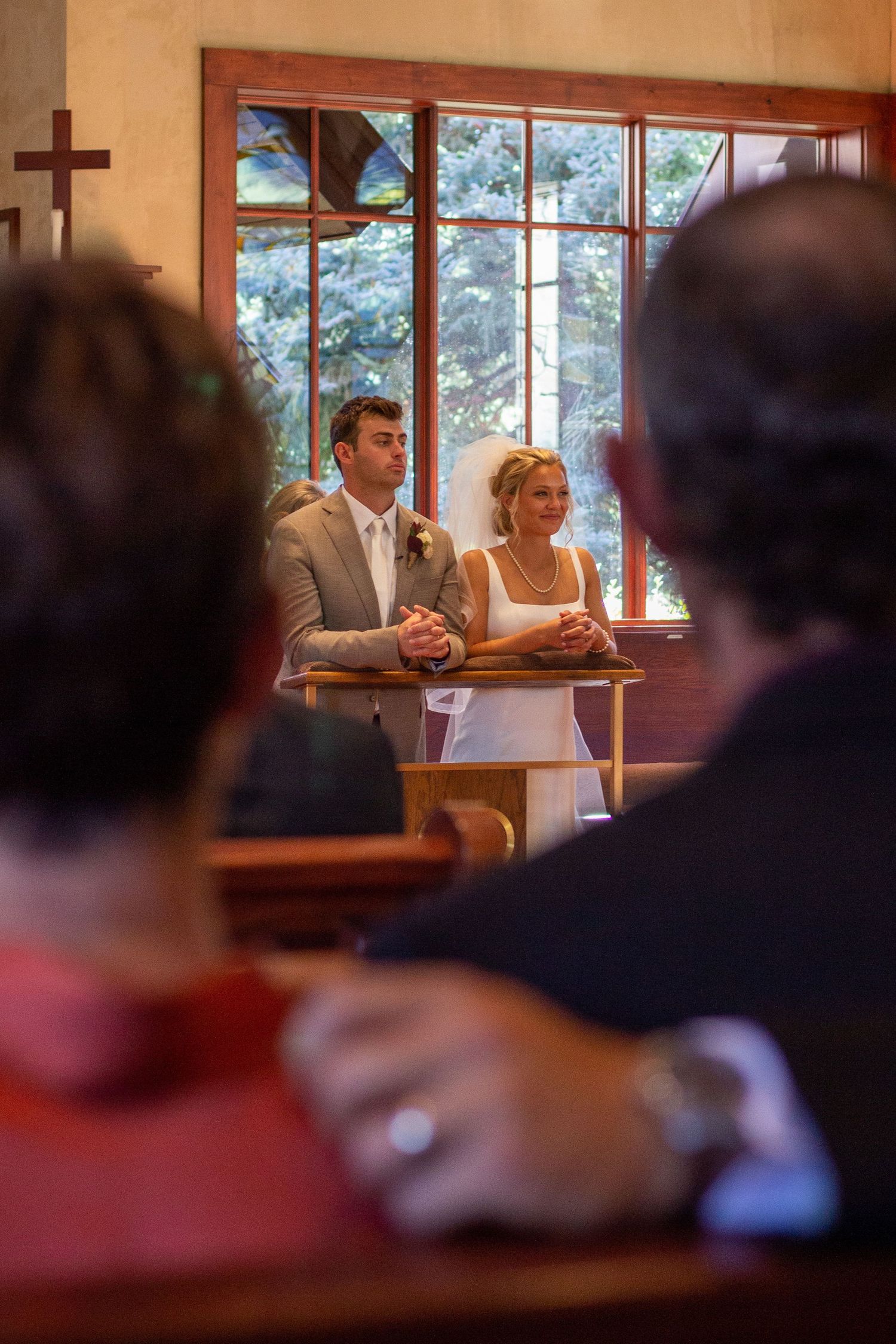Bride in white dress and groom in tan suit stand at a wooden altar during a catholic wedding ceremony as guests watch.