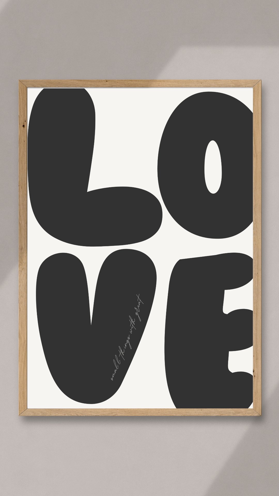 Framed black bubble-letter LOVE print with small script text on the V, in a light wood frame against a neutral wall.