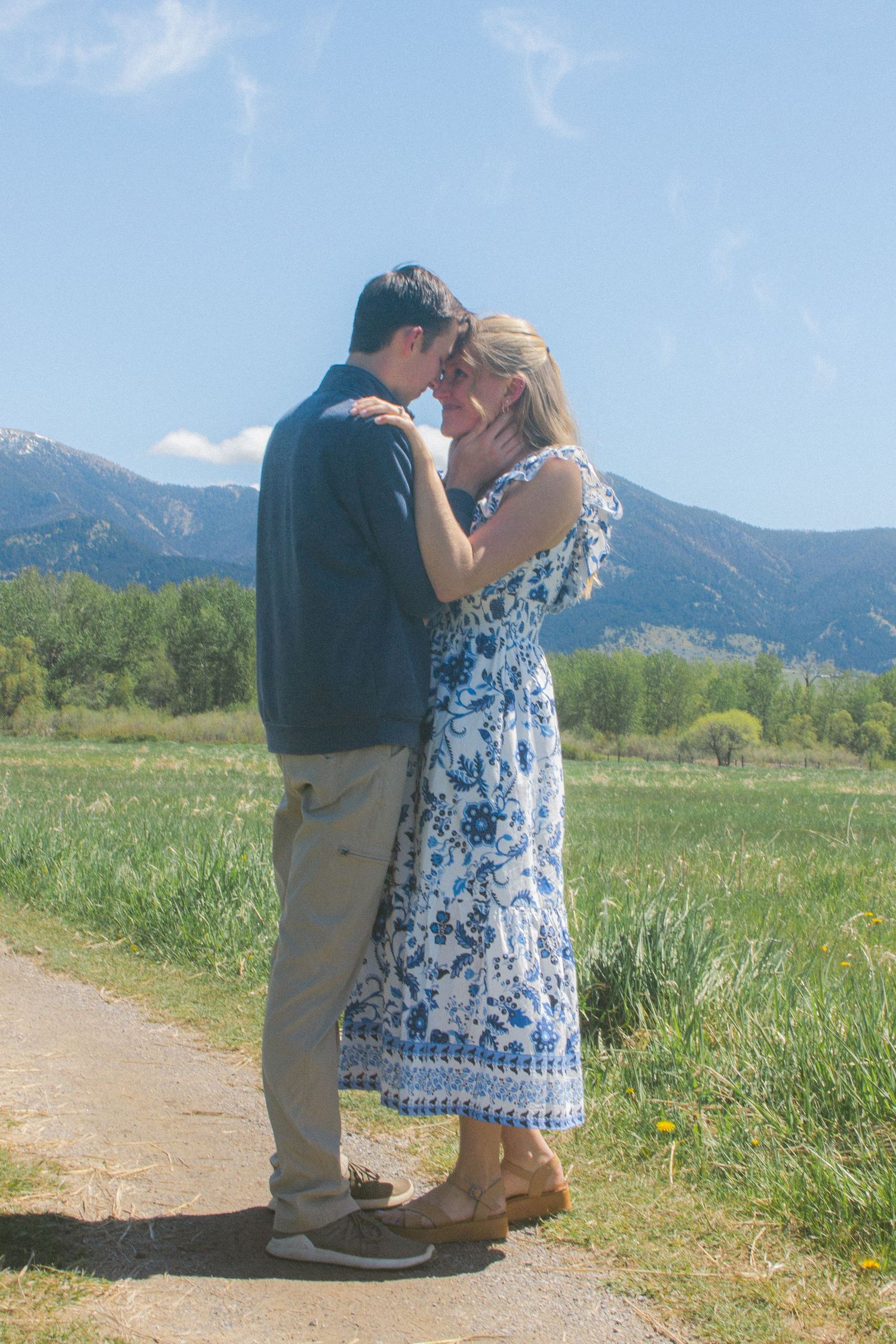 A couple embraces forehead to forehead on a dirt path in a green meadow with mountains and a bright blue sky behind them.