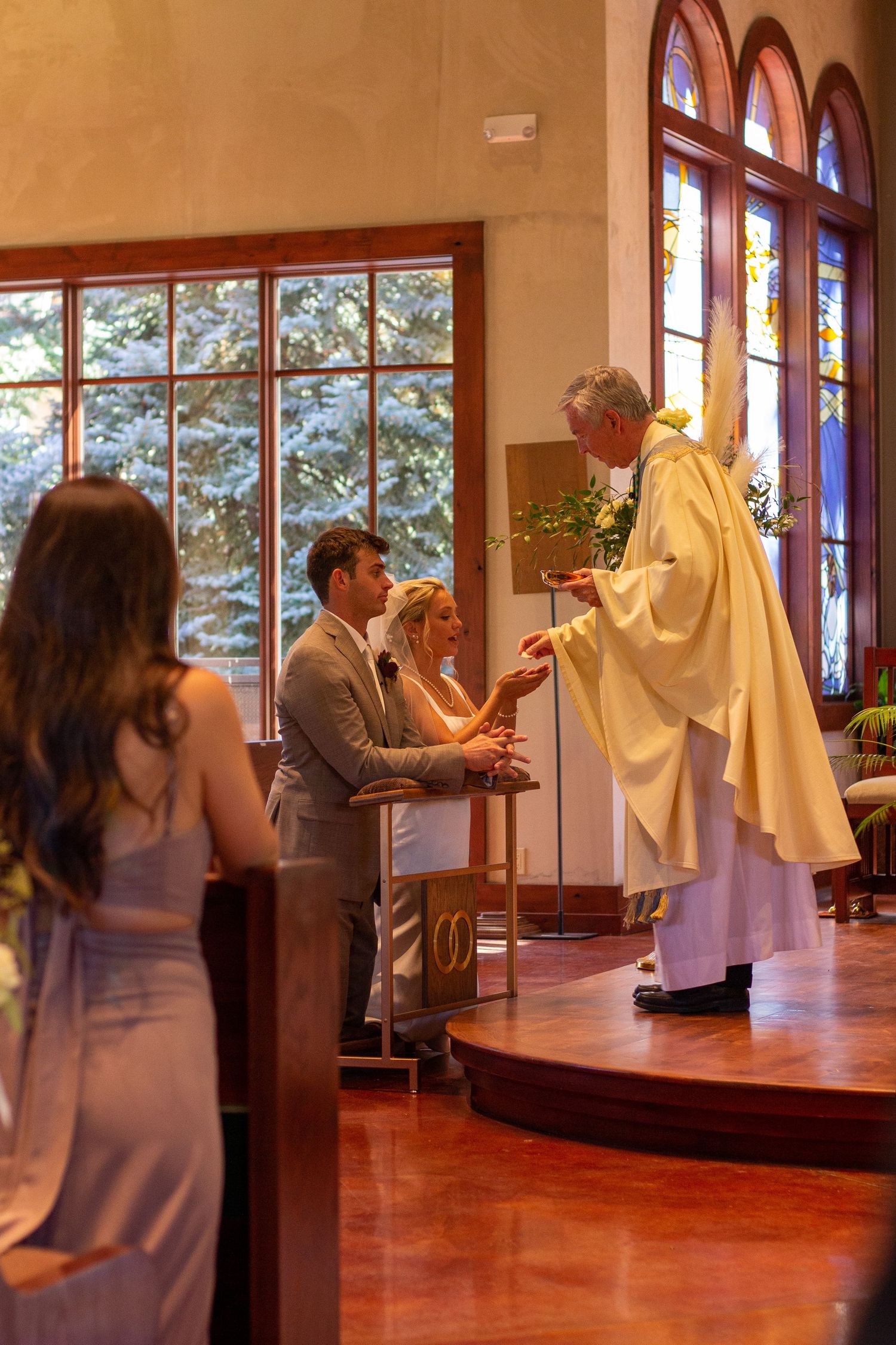 A priest in gold vestments blesses a bride and groom kneeling at the altar during a Catholic wedding ceremony as a bridesmaid watches.