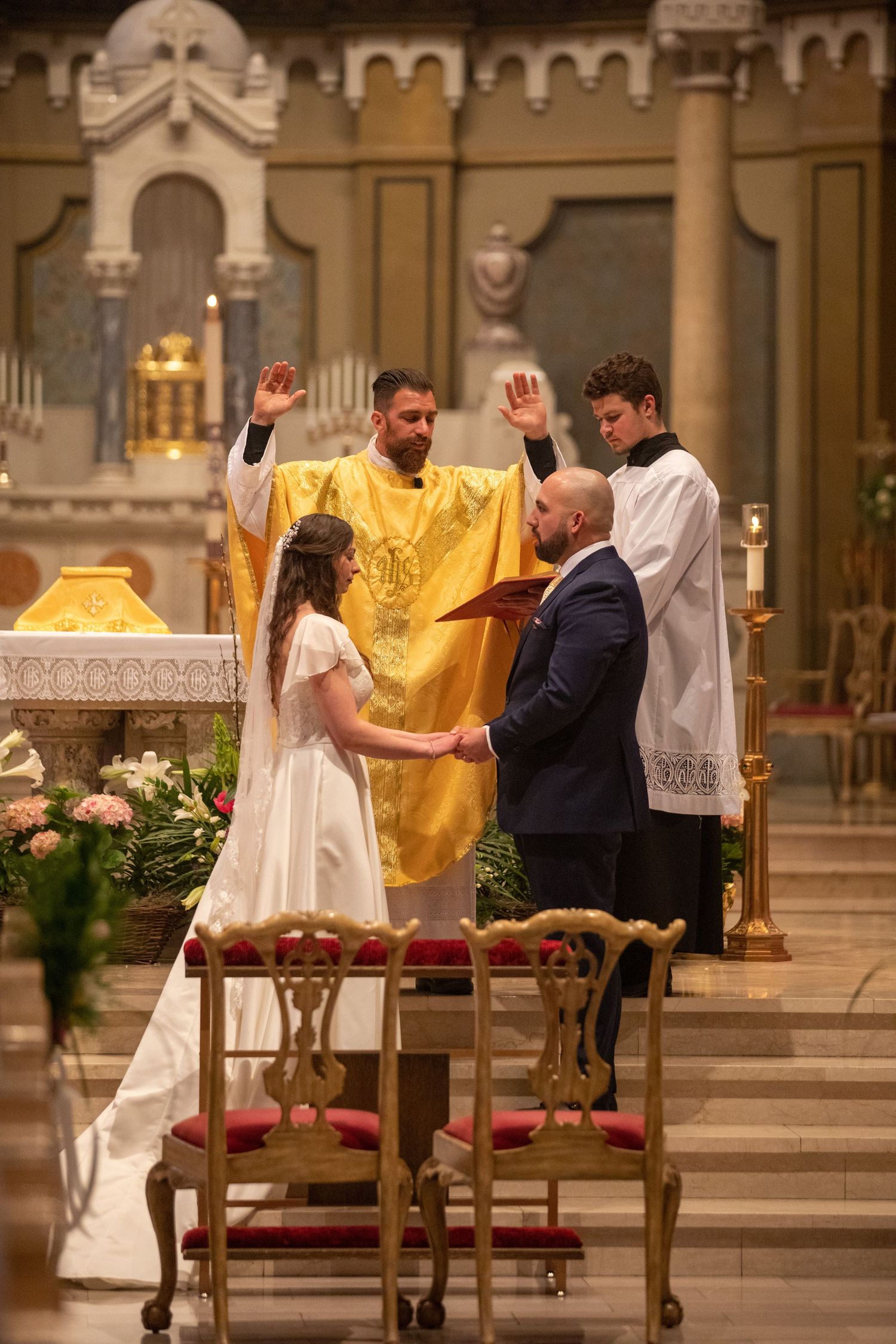 A bride and groom hold hands at the altar while a priest in gold vestments raises his hands in blessing and an altar server assists.