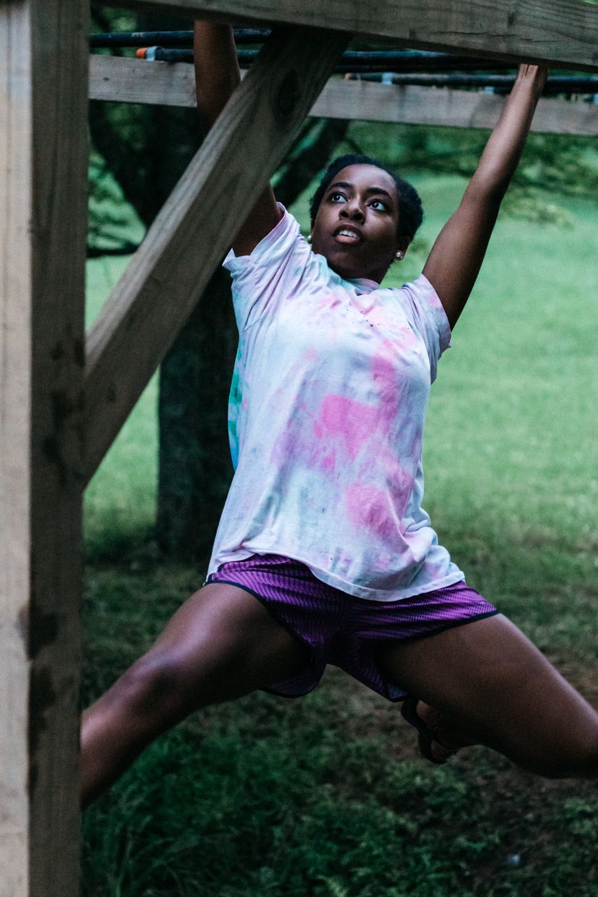 A young girl in a tie-dye shirt and purple shorts swings across wooden monkey bars outdoors on a green lawn.
