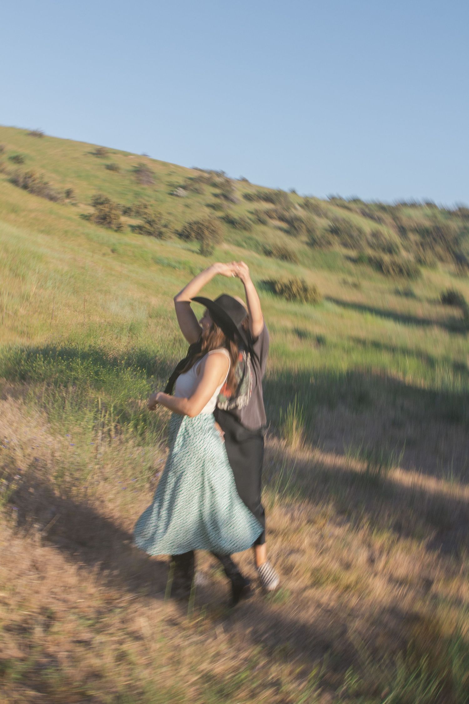 A woman in a black hat and green floral skirt twirls with raised arms on a sunlit grassy hillside.