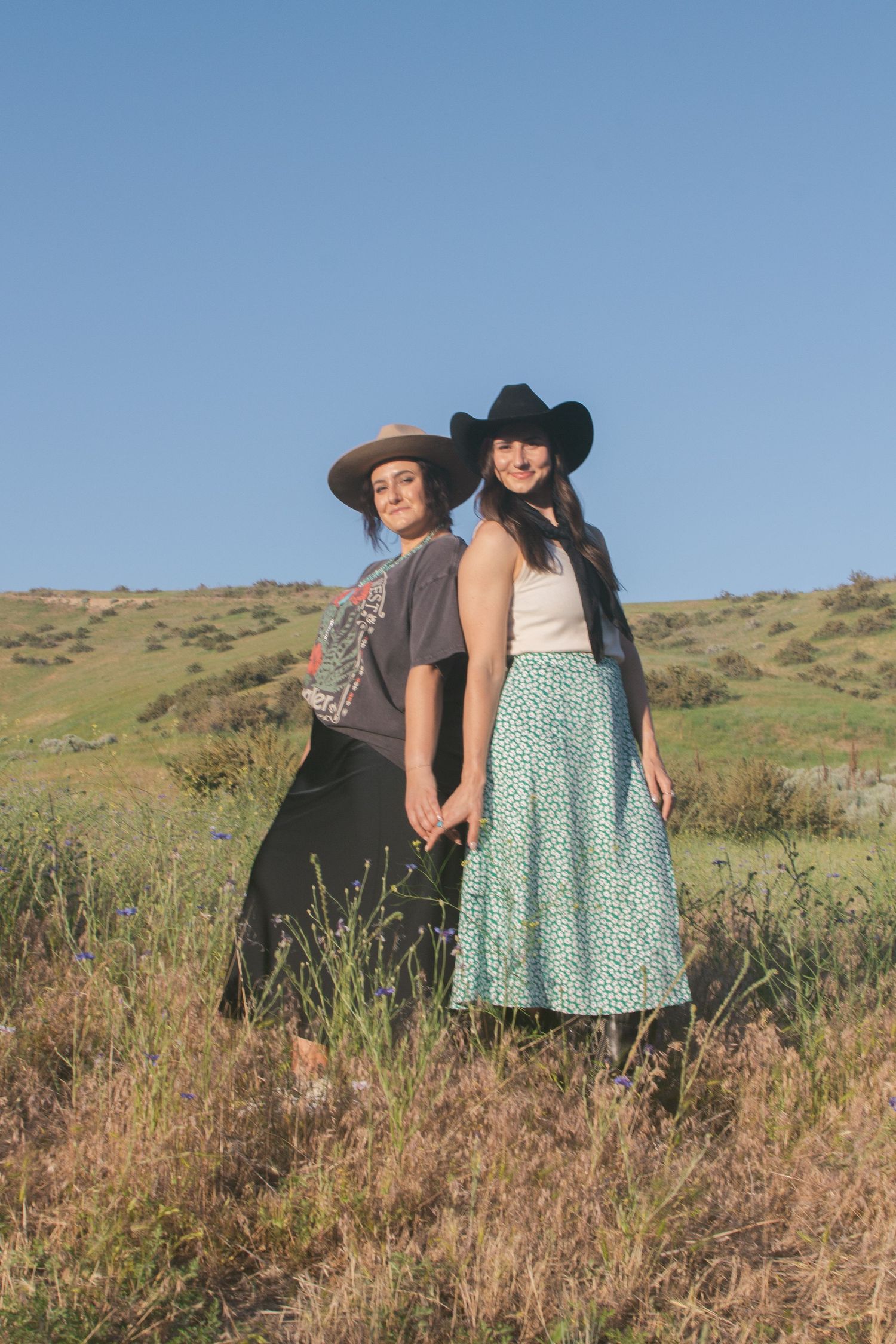 Two women in cowboy hats stand back to back in a wildflower field, one in a green floral skirt, the other in a graphic tee.