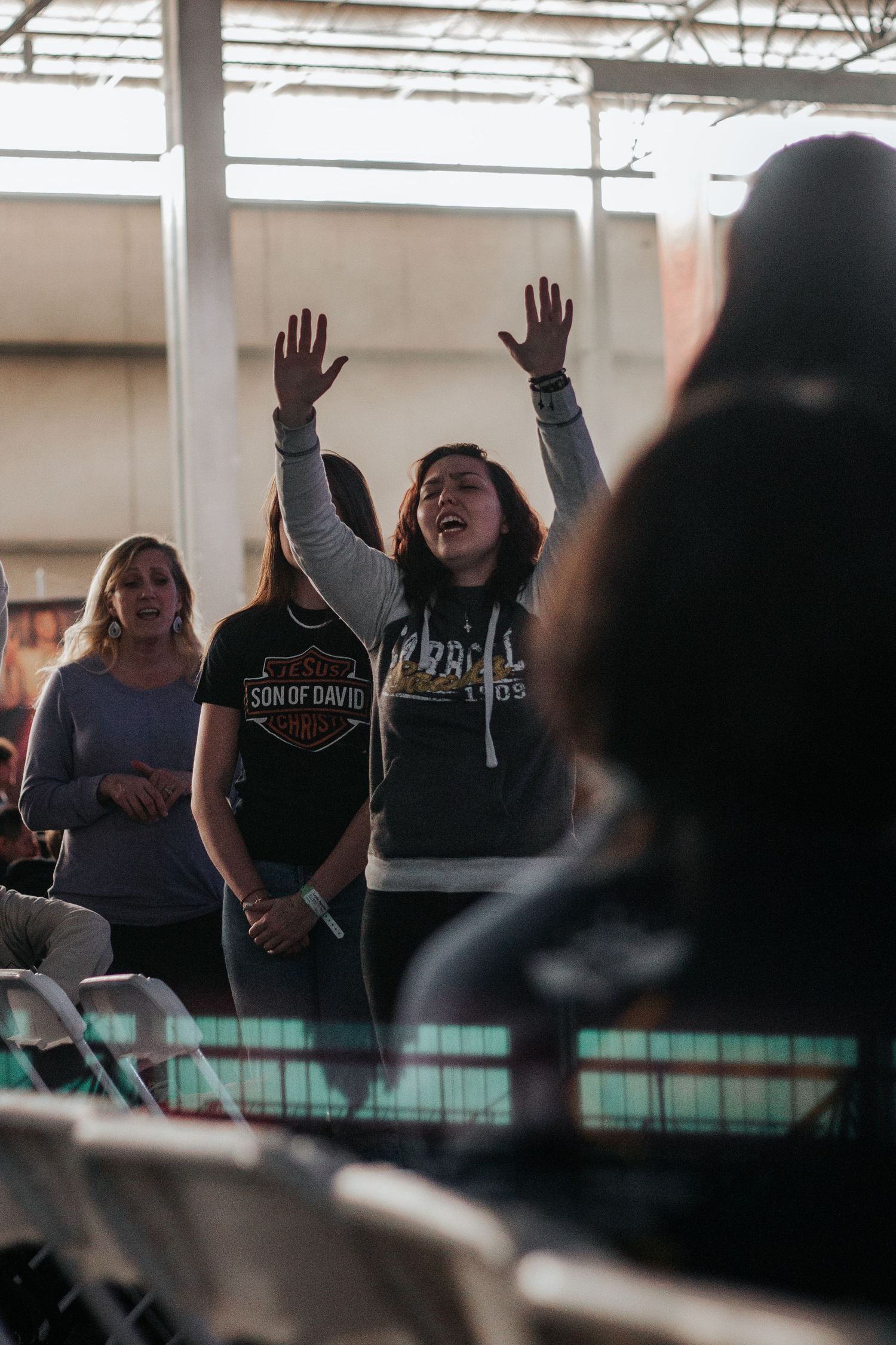 Young woman with raised hands sings with eyes closed during a worship service as people stand nearby in a large indoor venue.